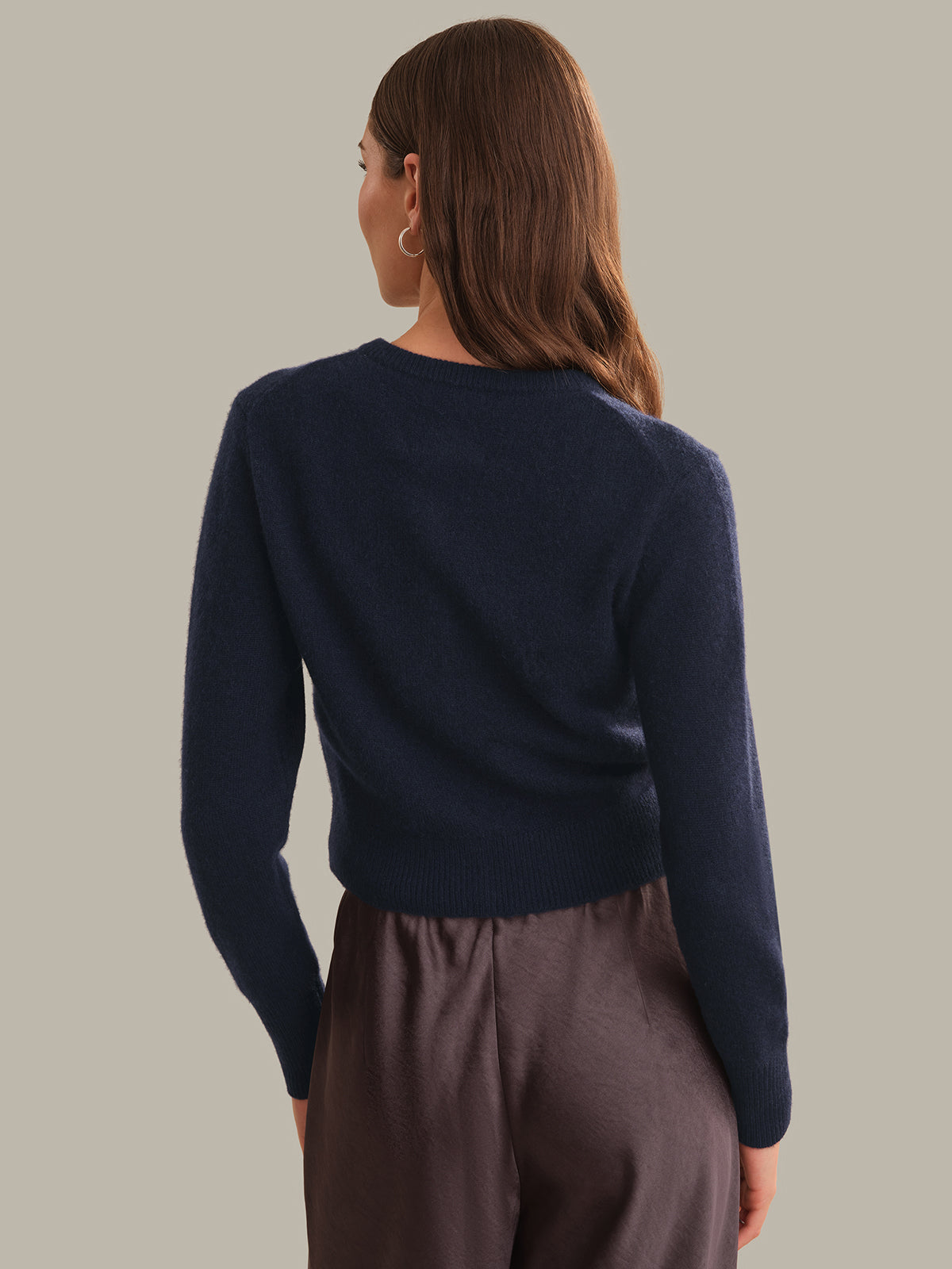 A woman shown from the back wearing a dark navy blue knit sweater and dark brown trousers, set against a solid light gray background.