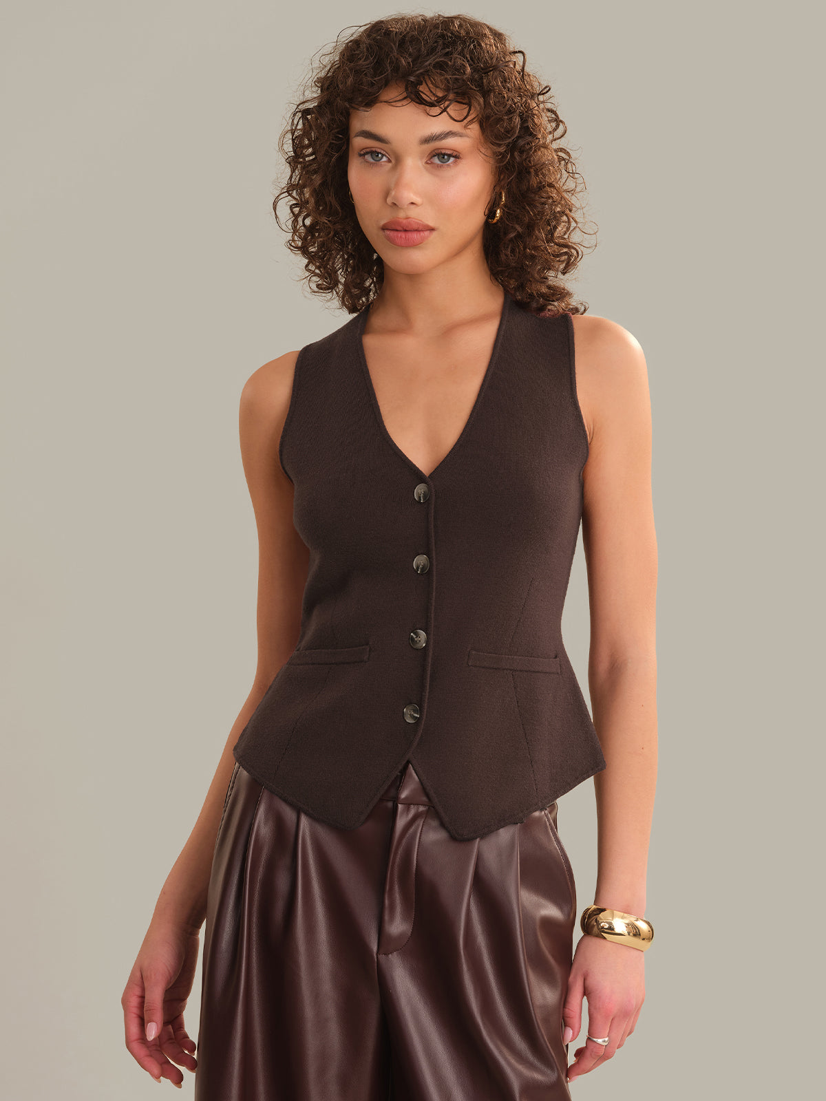 A model with curly brown hair wearing a sleeveless dark brown button-front vest and shiny dark brown satin pants, posed against a solid light gray studio background.