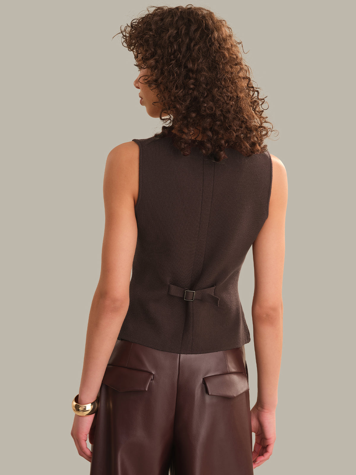 A woman shown from the back wearing a sleeveless dark brown fitted top with a small waist tie and dark brown leather-like pants, against a plain warm gray studio background.