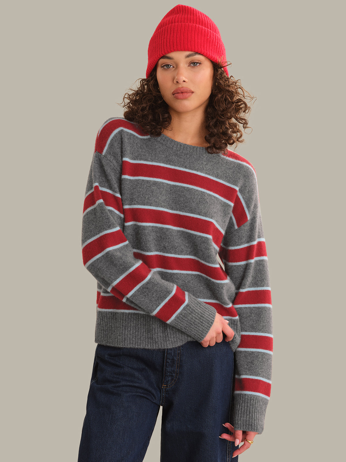 A woman wearing a gray knit sweater with horizontal red and white stripes, paired with dark blue jeans and a red beanie, against a solid light gray background.