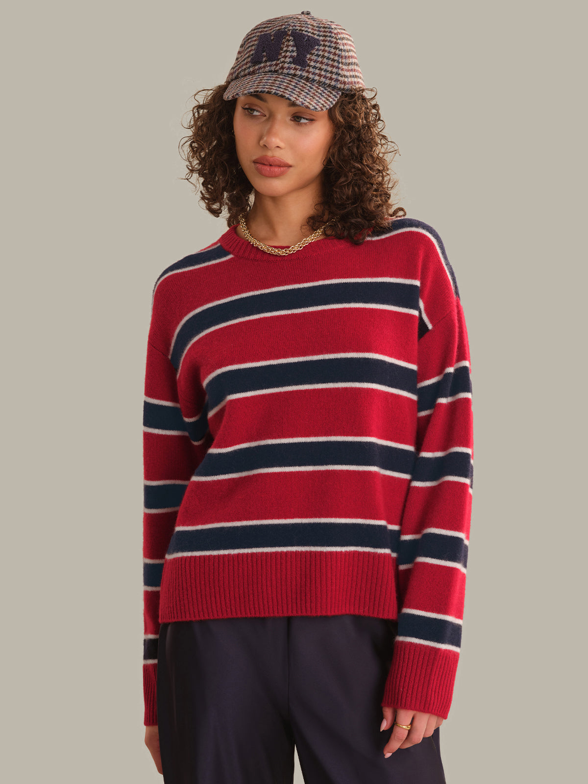 A woman wearing a red knit sweater with navy and cream horizontal stripes, paired with dark trousers and a plaid cap, against a solid gray background.