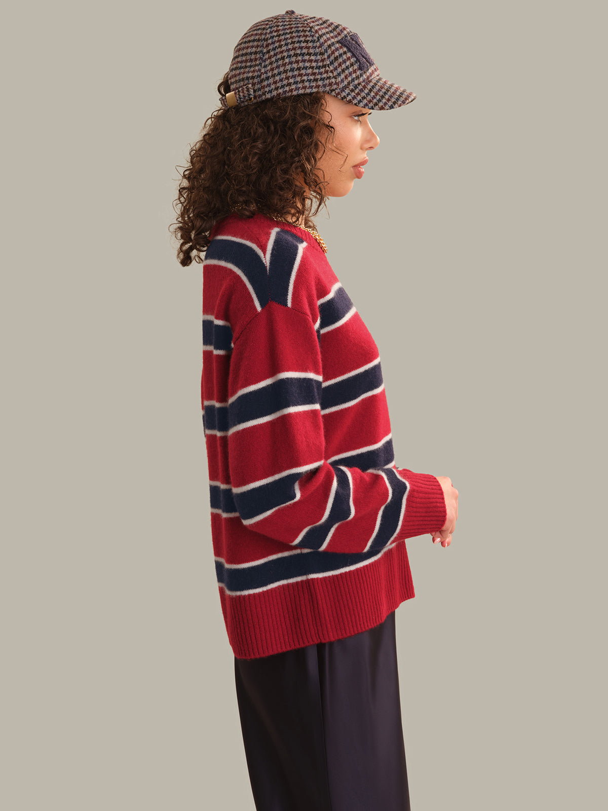 A woman shown in side profile wearing a red, navy, and white striped knit sweater, a brown and blue checkered cap, and dark pants, set against a solid light gray background.