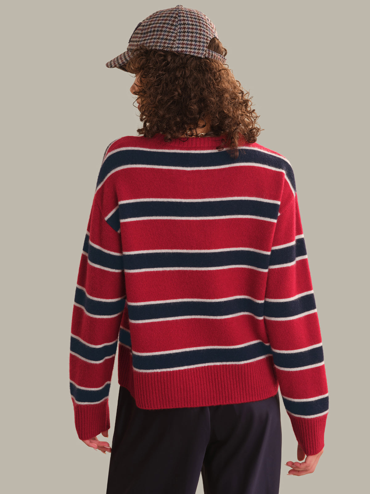 A person shown from the back wearing a red knit sweater with navy and white horizontal stripes, paired with dark pants and a plaid cap, against a plain light gray background.