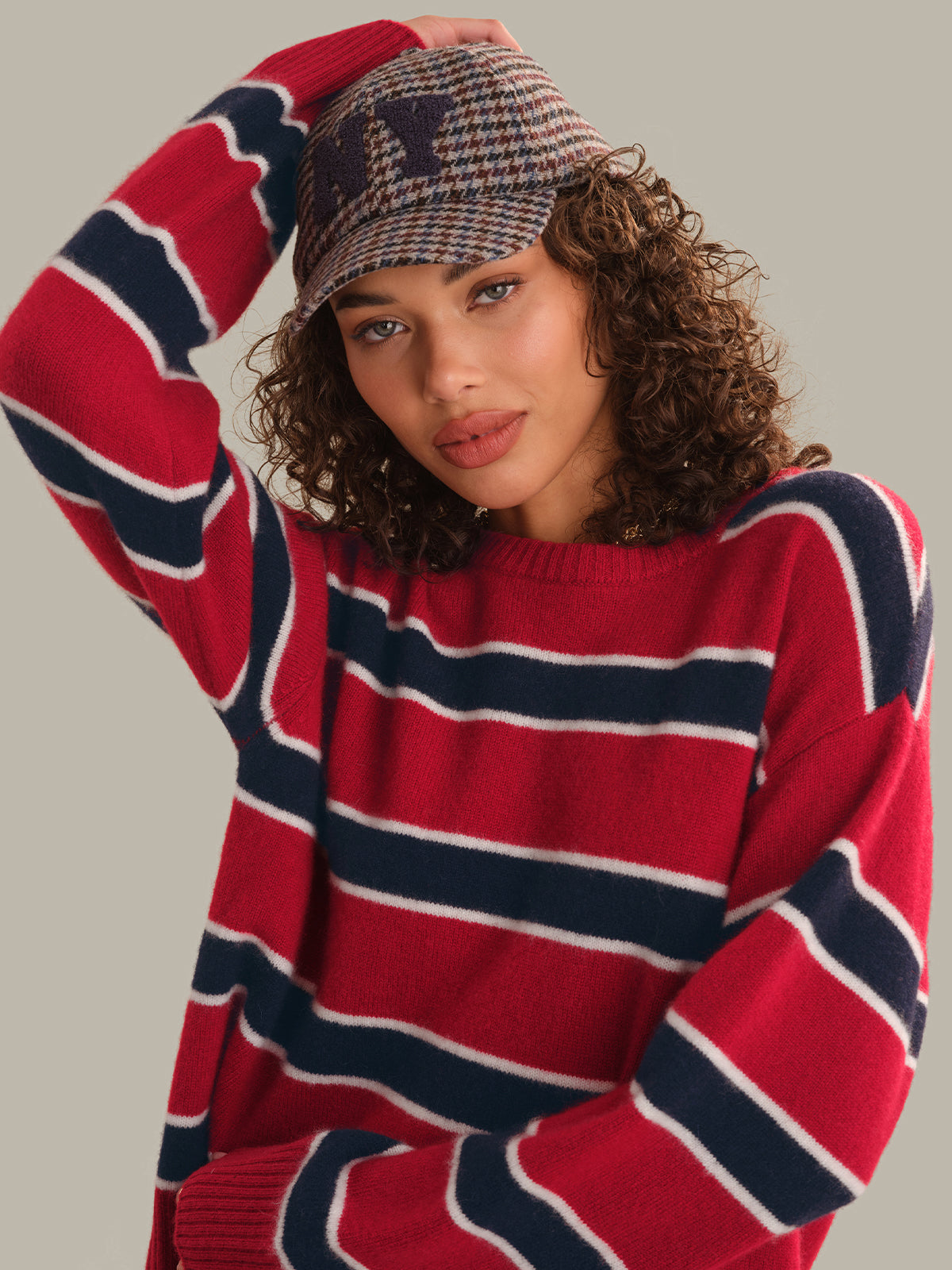 A woman wearing a red, navy, and white striped knit sweater and a matching patterned knit cap, posed against a solid light gray background.
