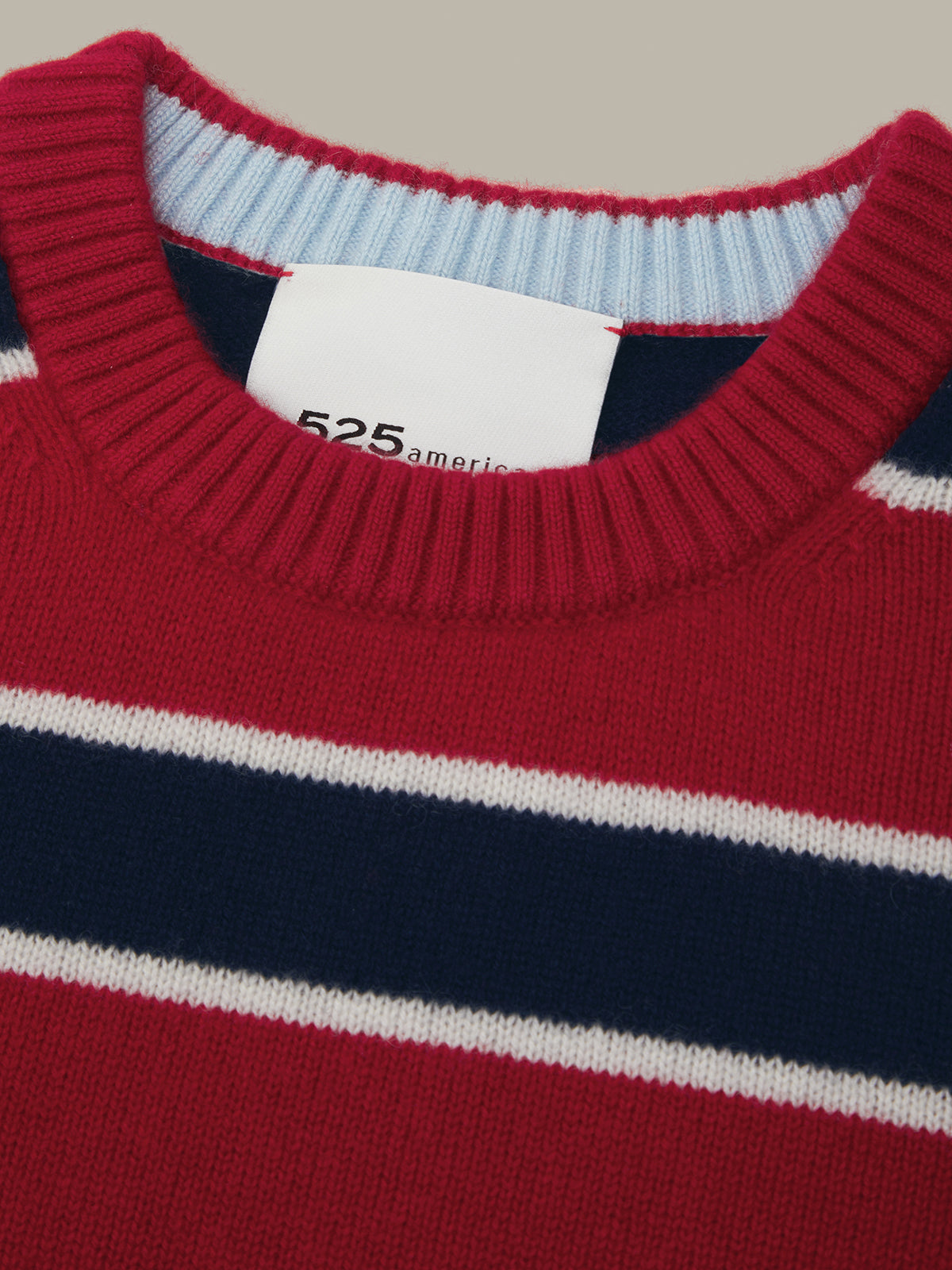 Close-up of a striped knit sweater showing a red ribbed crew neck, a white neck label reading '525 america', and horizontal navy and white stripes.
