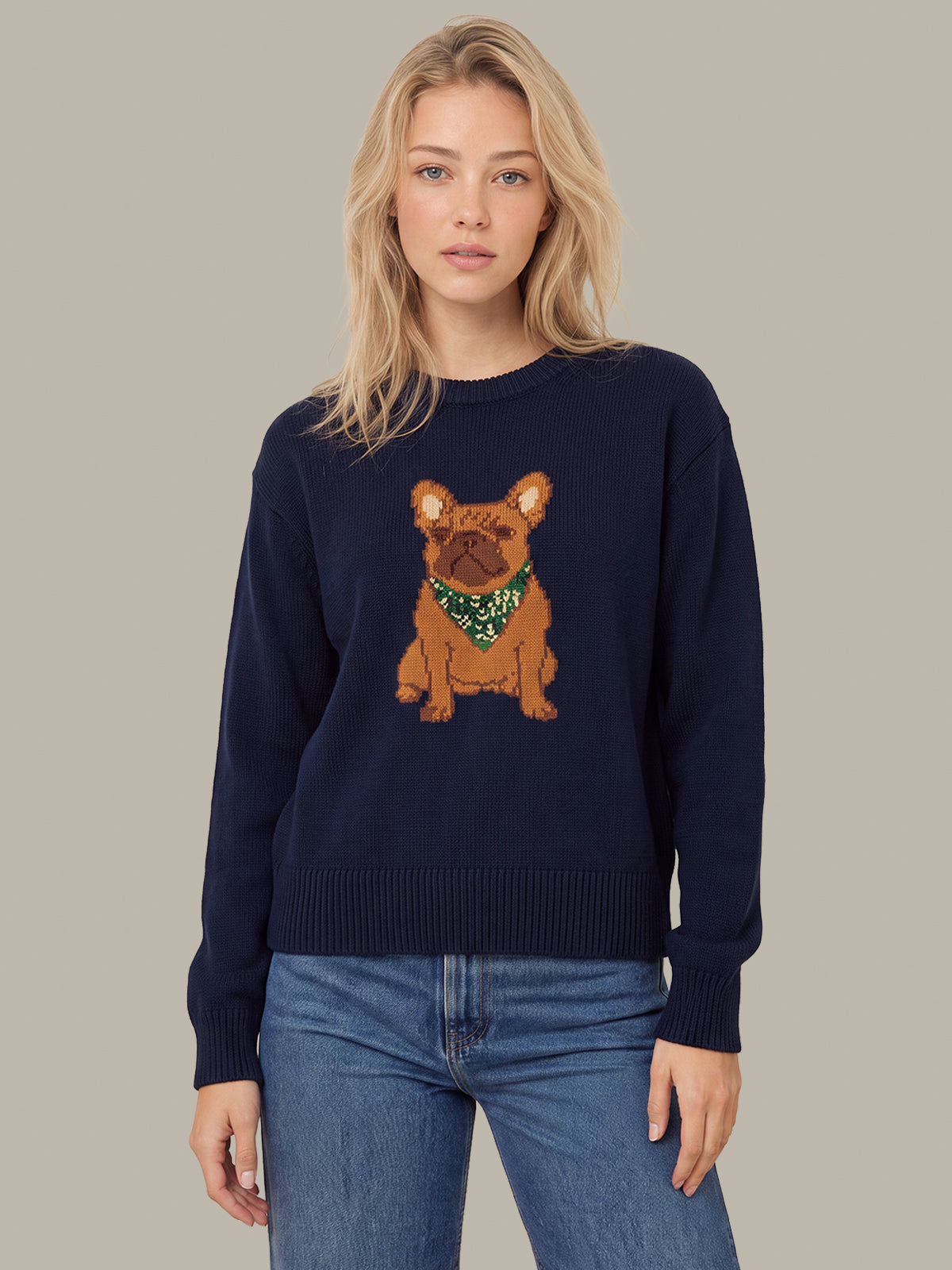 A woman wearing a navy blue crewneck sweater with a graphic of a brown French Bulldog wearing a green patterned bandana, paired with blue jeans, against a solid taupe background.