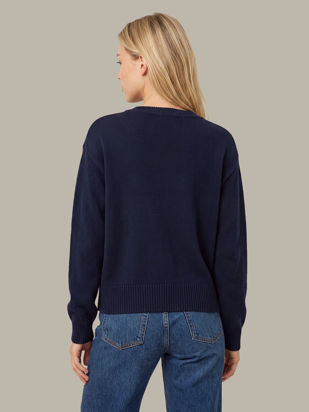 A woman shown from the back wearing a dark navy blue knit sweater and blue jeans, standing against a solid light gray background.