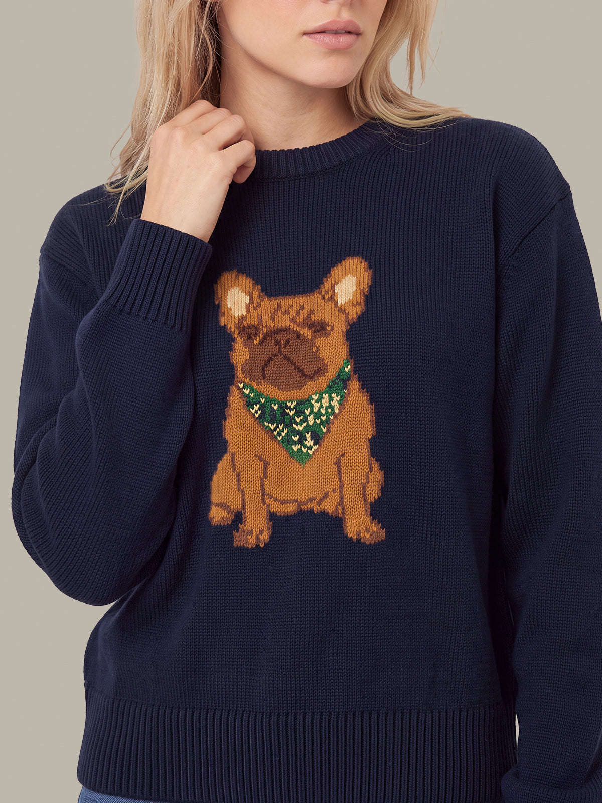 A woman wearing a navy blue ribbed knit sweater featuring a knitted French Bulldog graphic with a green bandana on the chest, set against a solid beige background.