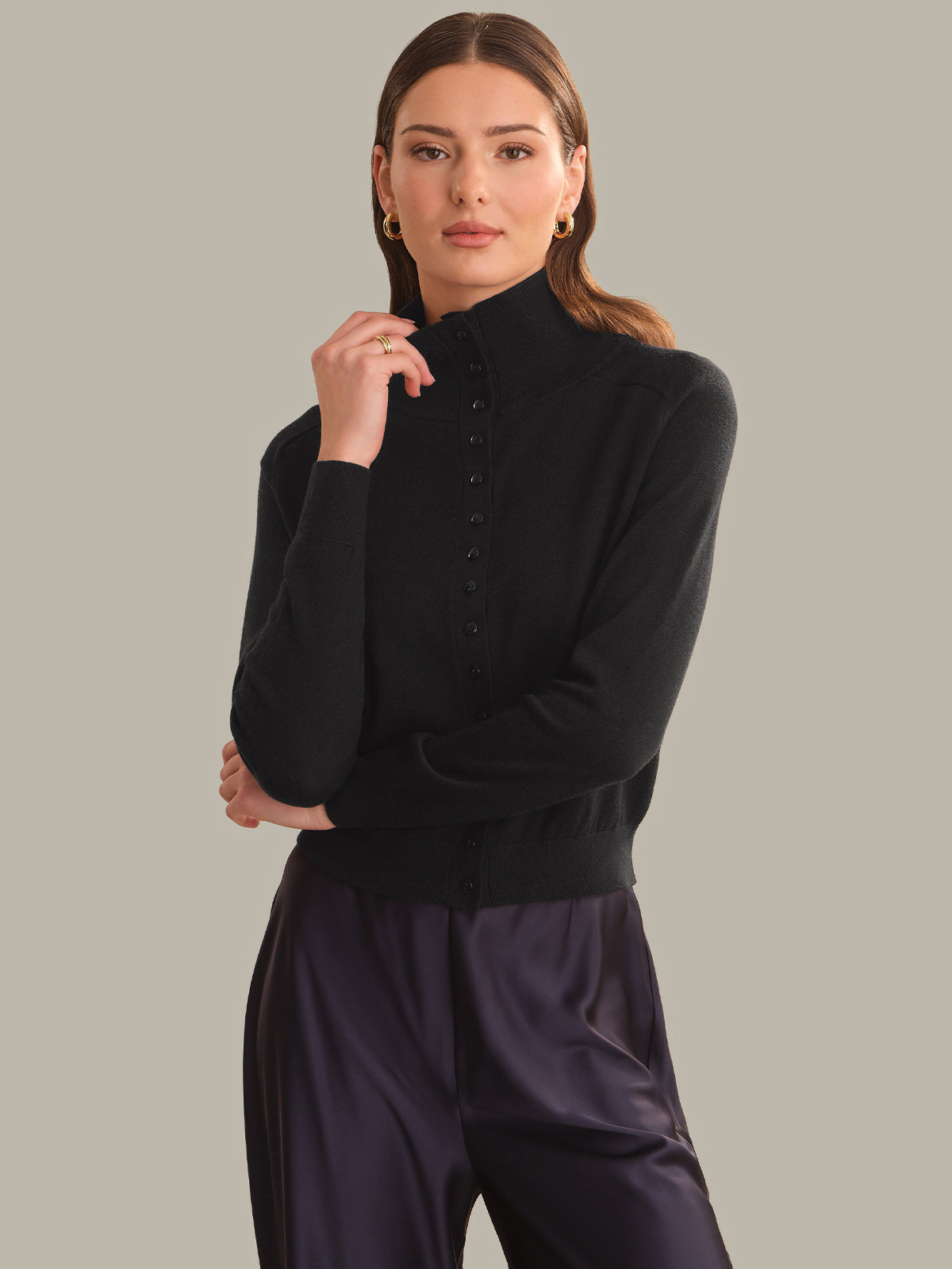 A woman modeling a black mock-neck knit top with a buttoned front and dark purple satin trousers, set against a solid light gray studio background.