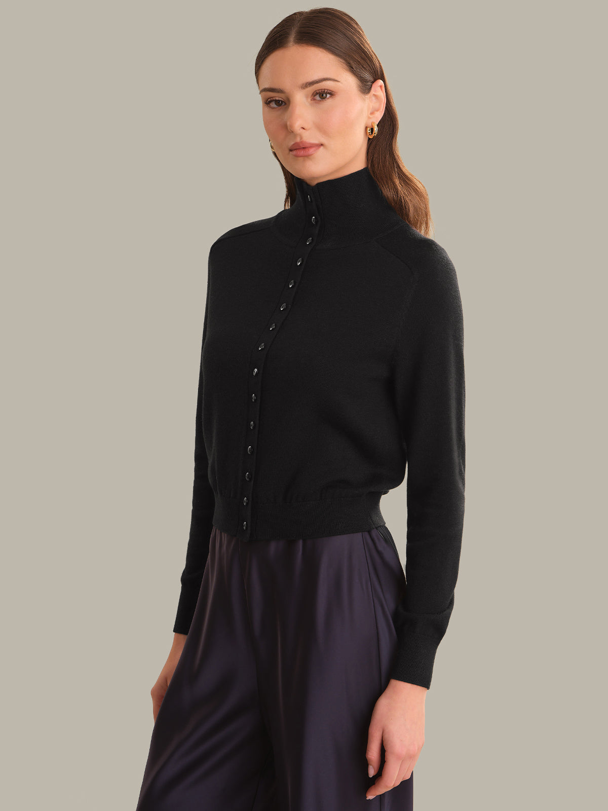 A woman modeling a black long-sleeve mock neck knit cardigan with decorative buttons down the front, paired with dark purple satin trousers, against a solid gray studio background.