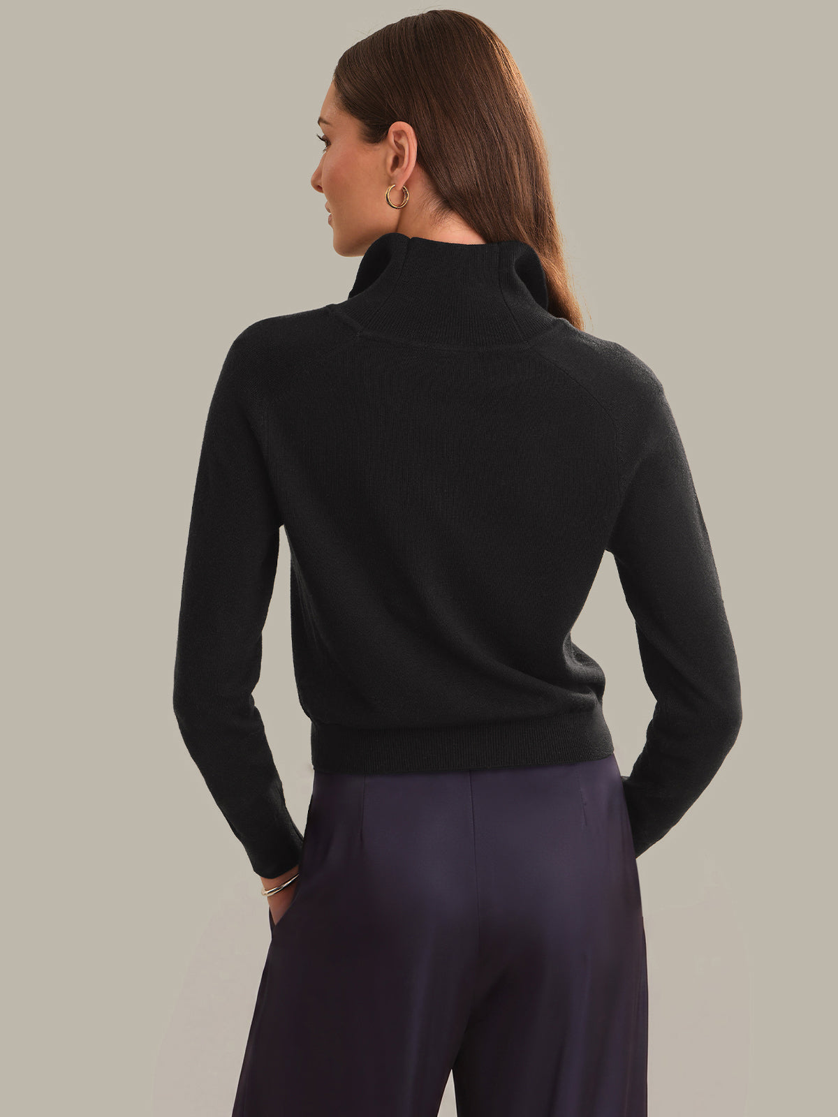 A woman shown from the back wearing a black ribbed mock neck sweater and dark purple trousers, set against a solid light gray studio background.