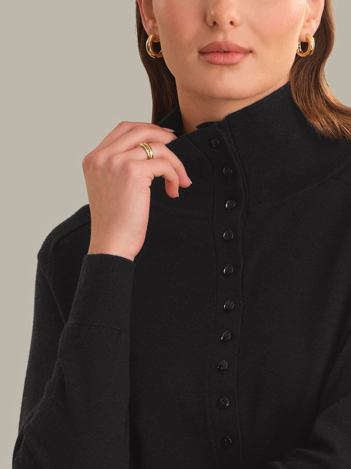 Close-up of a model wearing a black knit mock-neck button-up top, featuring a high collar and black buttons down the front, set against a solid warm gray background.