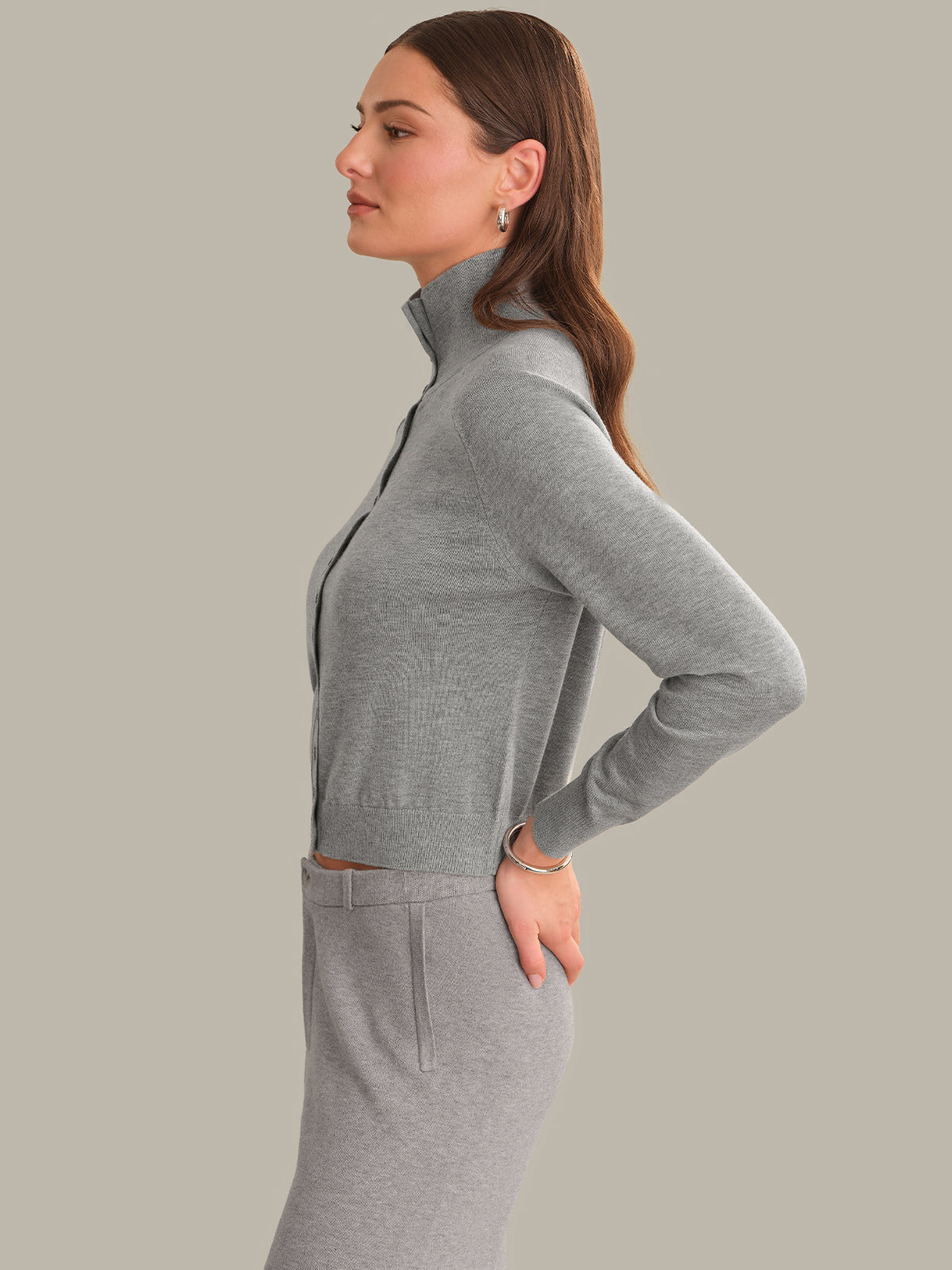 A woman in side profile wearing a light gray ribbed mock-neck long-sleeve top and matching gray skirt against a solid light gray background.