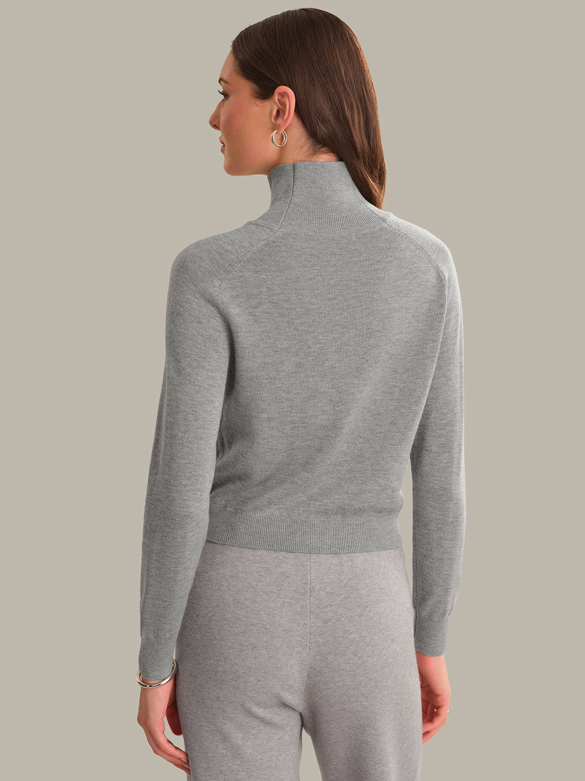 A woman shown from the back wearing a light gray ribbed mock neck sweater and matching gray pants against a solid beige background.