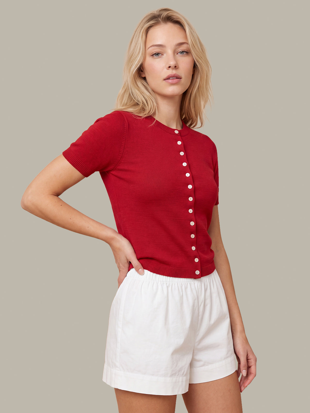 A woman modeling a short-sleeve red knit top with a vertical row of white buttons down the front, paired with white shorts, against a solid taupe background.