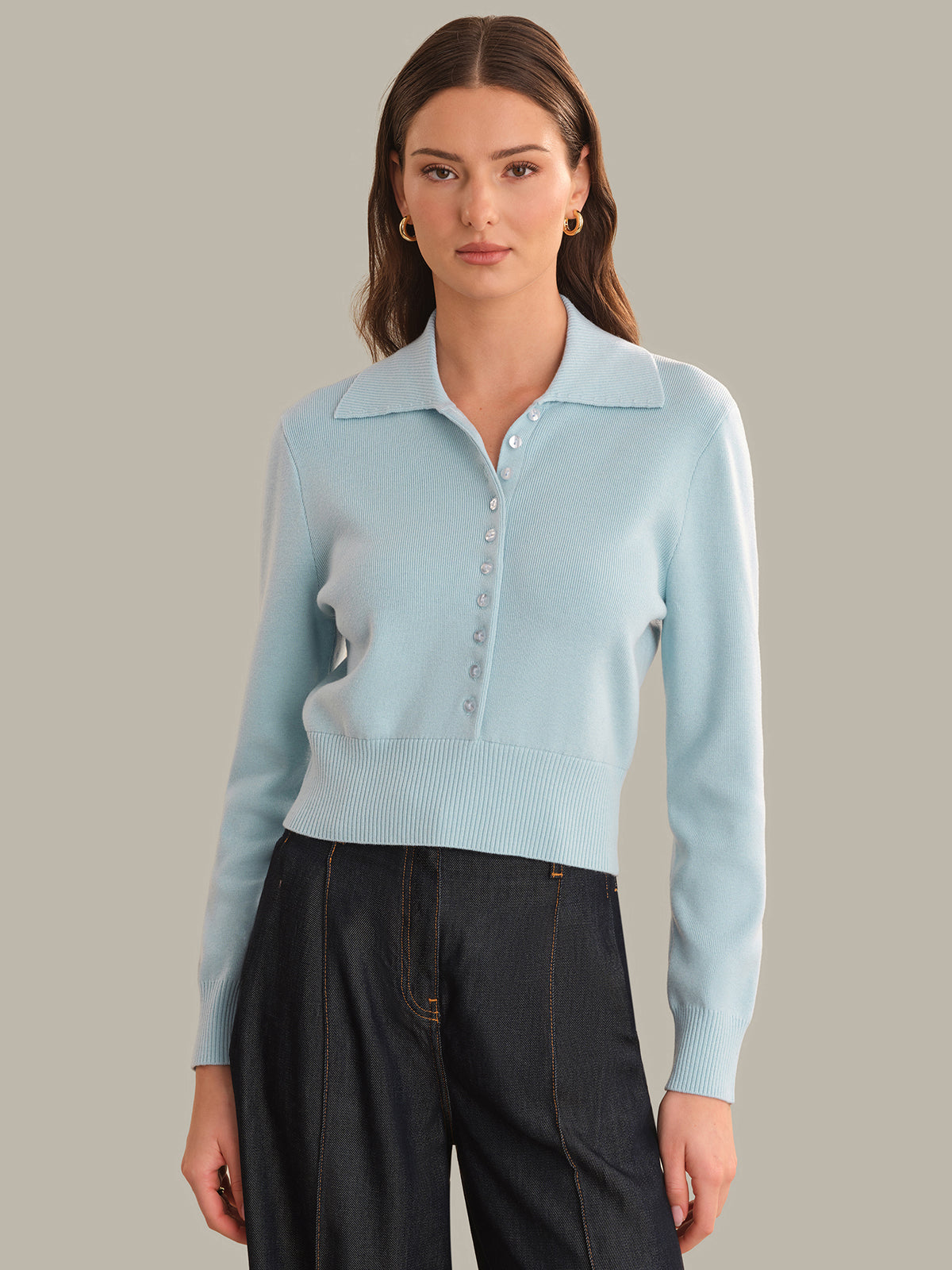 A woman modeling a light blue knit polo sweater with a buttoned placket, paired with dark charcoal pants, against a solid warm gray background.