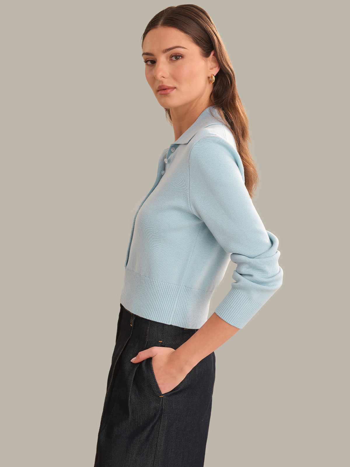 A woman wearing a light blue ribbed knit polo sweater and dark charcoal pants, shown in a side profile against a solid warm gray studio background.