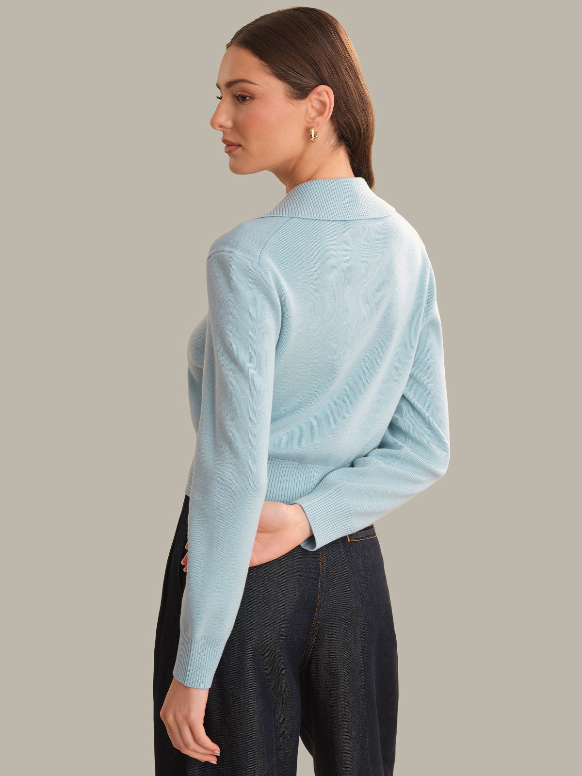 A woman shown from the back wearing a light blue mock neck knit sweater and dark charcoal pants, set against a solid warm gray studio background.