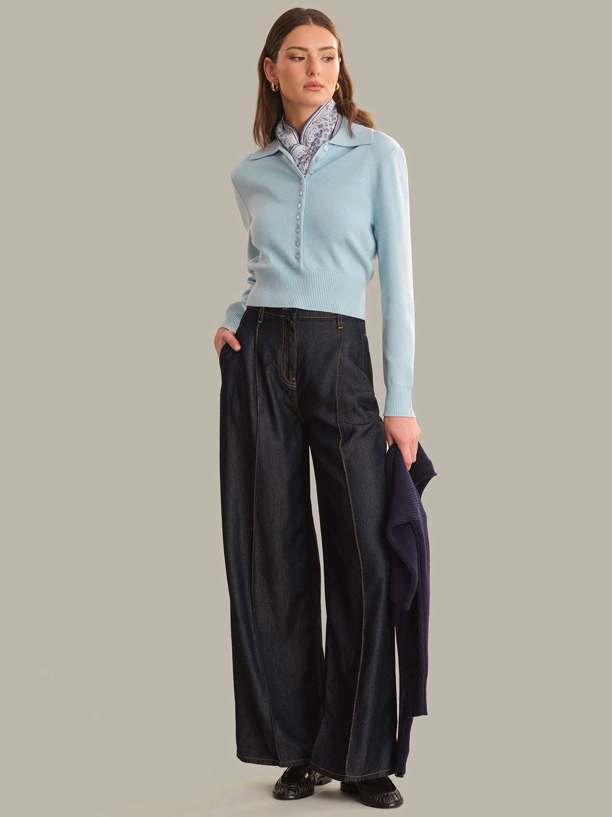 A woman modeling a light blue knit polo sweater with a buttoned collar, paired with wide-leg dark trousers and holding a matching light blue sweater in one hand, against a solid beige background.