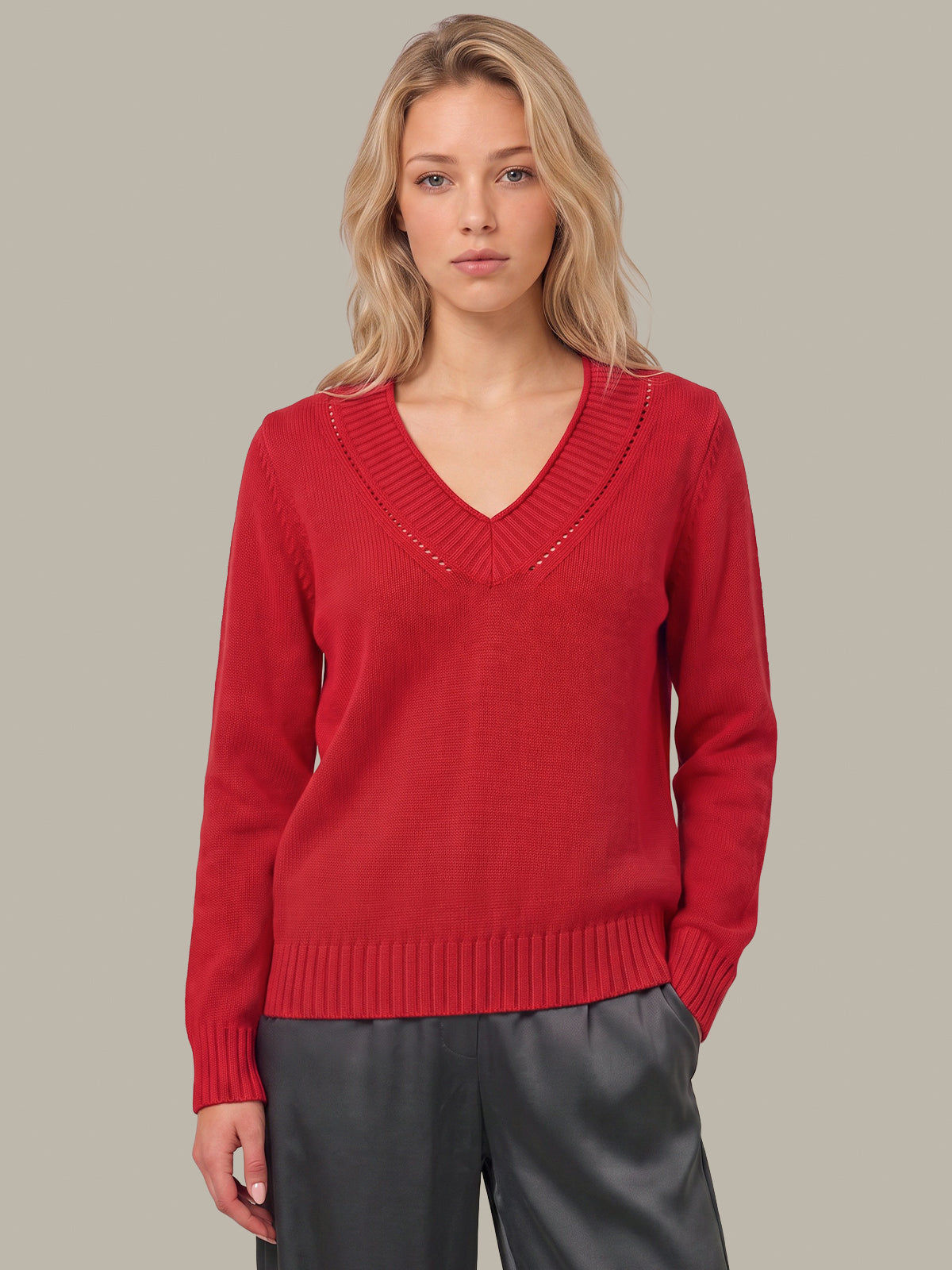 A woman wearing a red knit V-neck sweater with decorative openwork detailing around the neckline, paired with dark gray pants, against a solid light gray studio background.