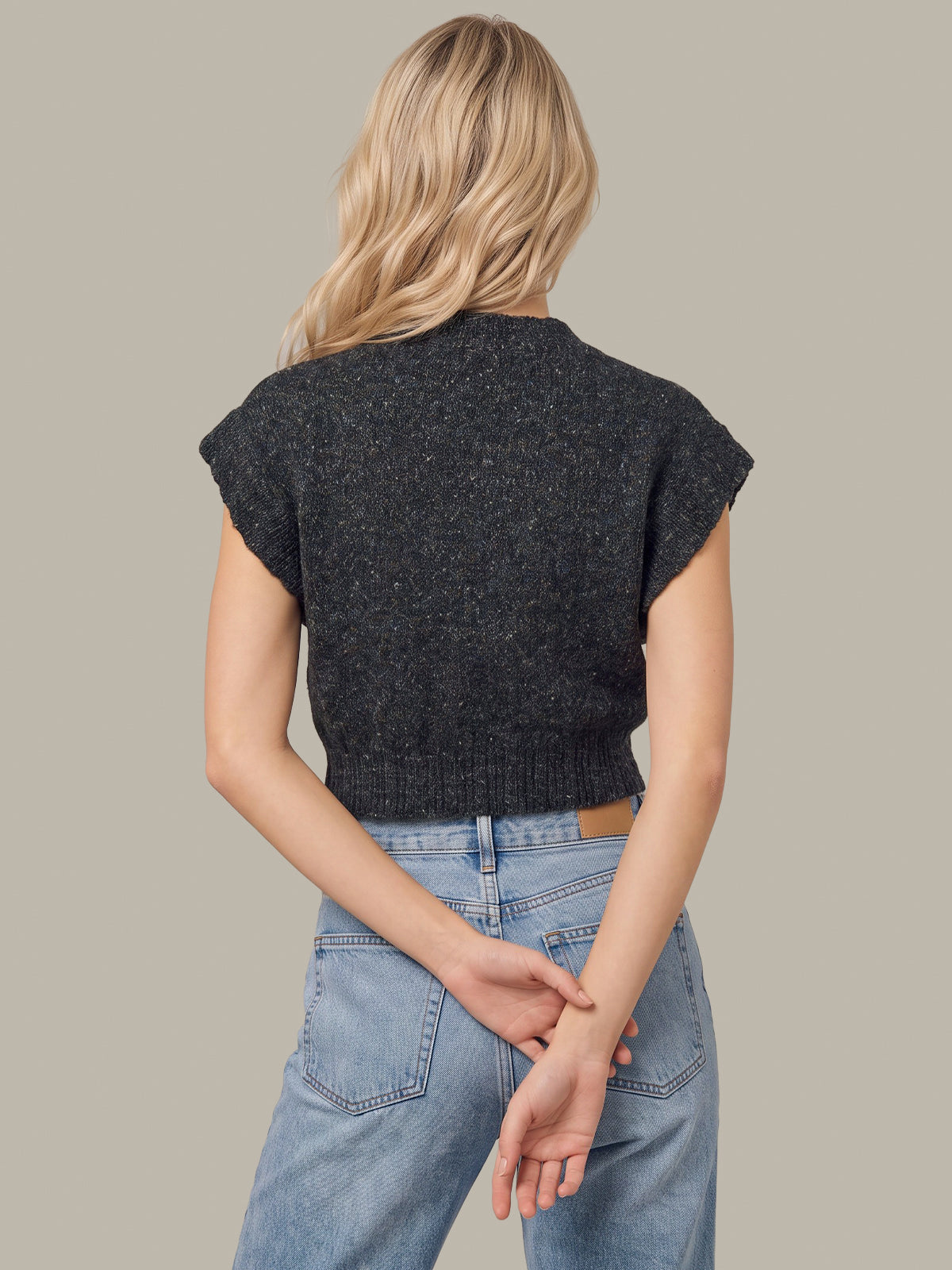 A woman shown from behind wearing a dark charcoal gray cropped knit sweater with cap sleeves and light blue jeans, set against a solid warm gray background.