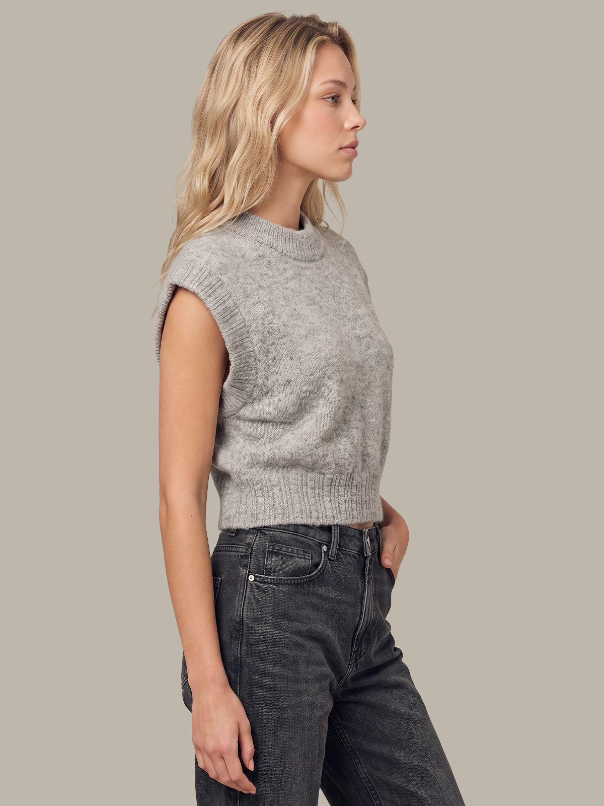 A woman in profile wearing a light gray sleeveless ribbed knit sweater vest and dark gray jeans, set against a solid light gray studio background.