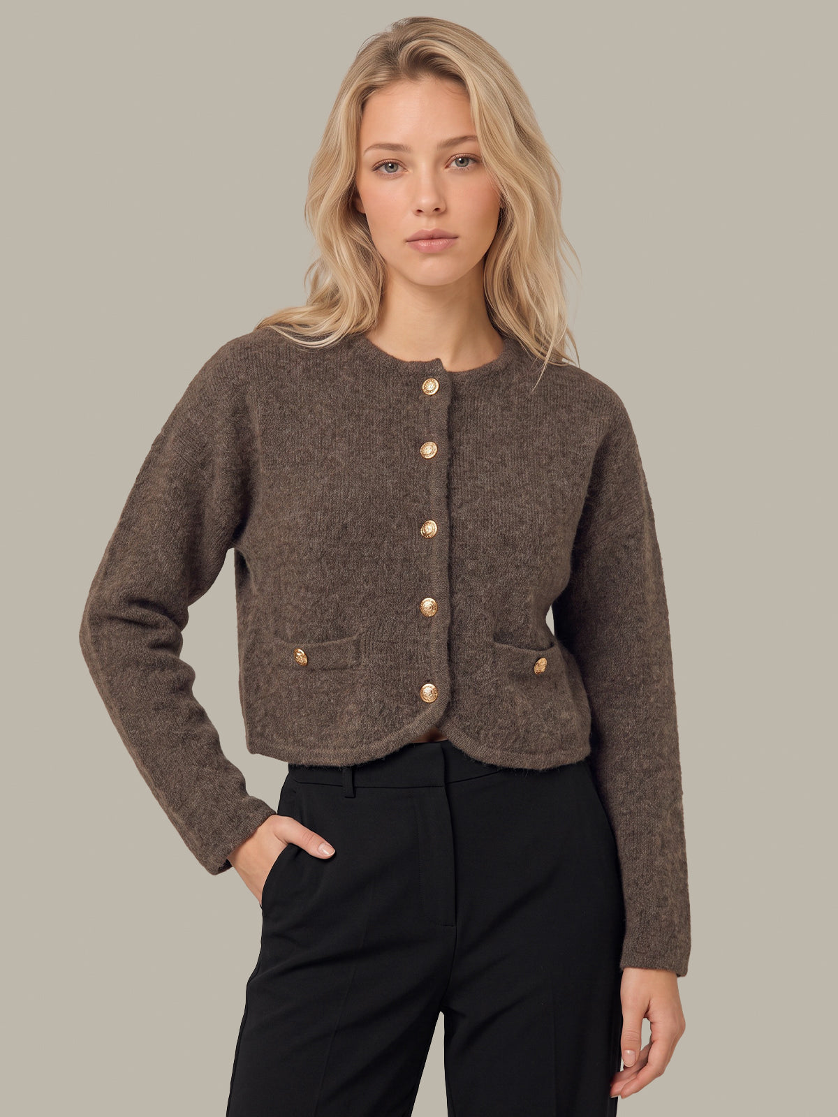 A blonde woman modeling a cropped, textured brown cardigan with gold-tone buttons and flap pockets, paired with black trousers, against a solid light gray background.
