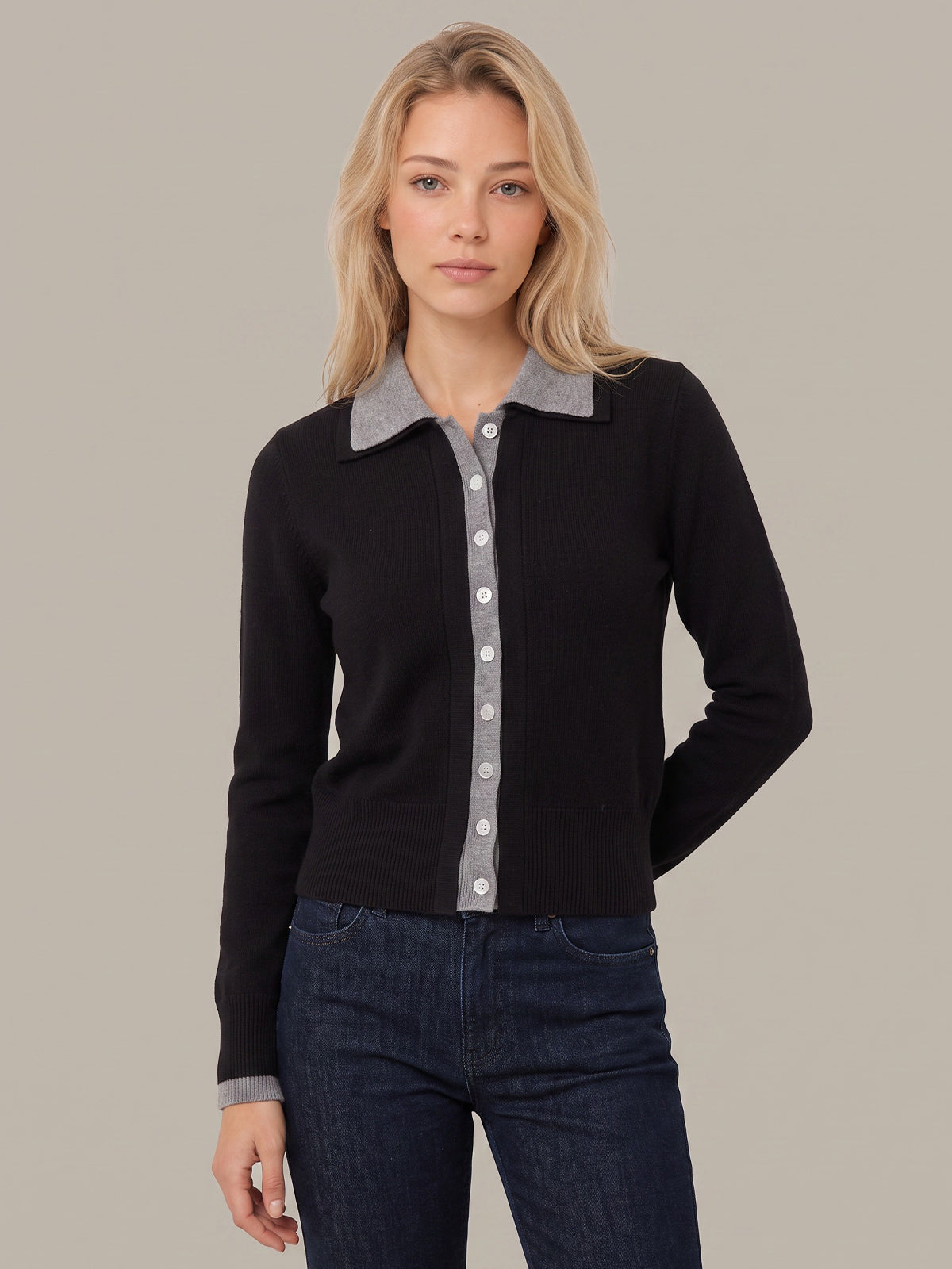 A woman modeling a black long-sleeve knit cardigan with a contrasting light gray button-front placket and matching light gray collar, paired with dark blue jeans against a solid light gray studio background.