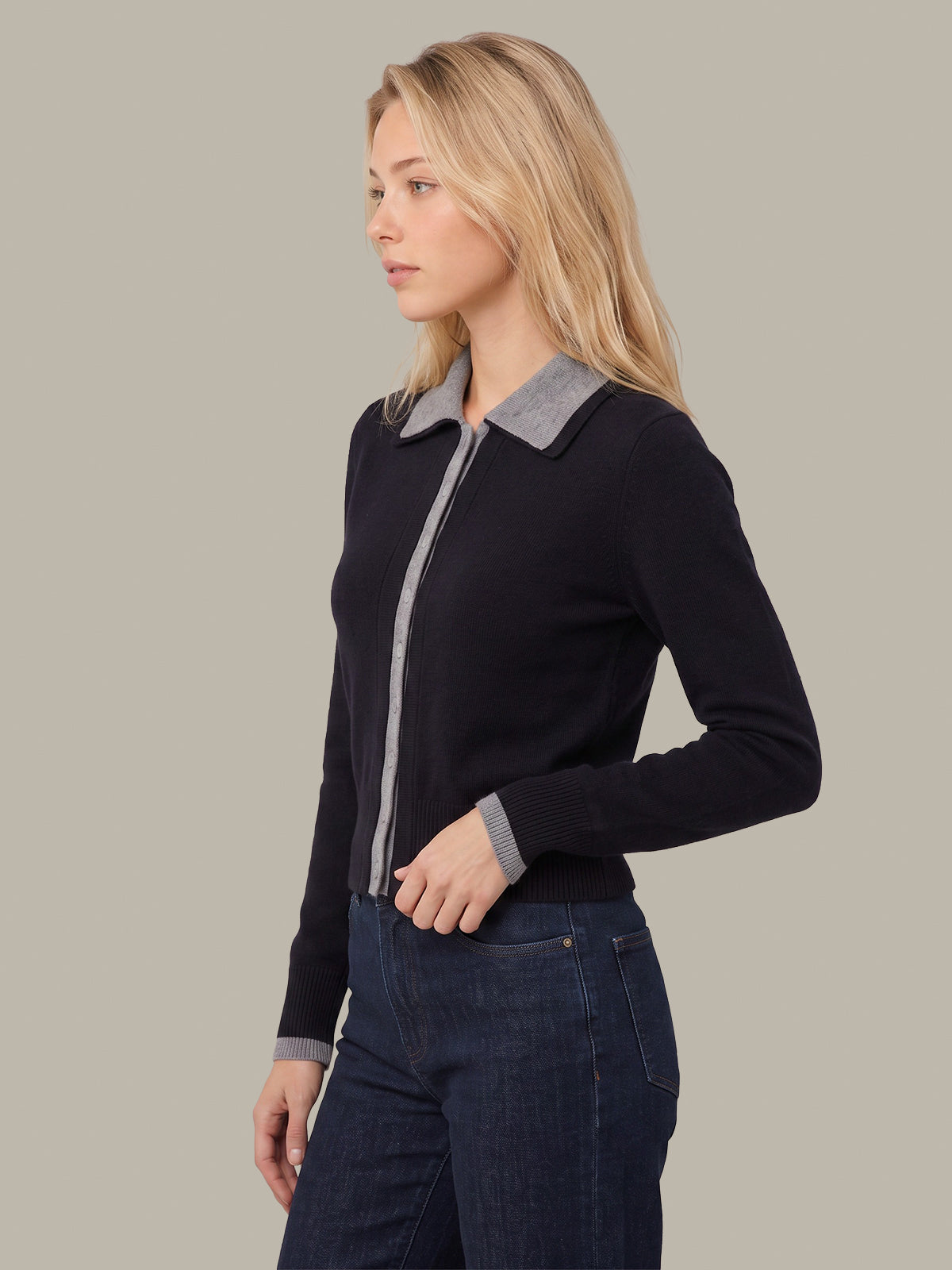 A woman shown in side profile wearing a dark navy knit zip-front cardigan with a contrasting light gray collar and cuffs, paired with dark blue jeans, against a solid light gray studio background.