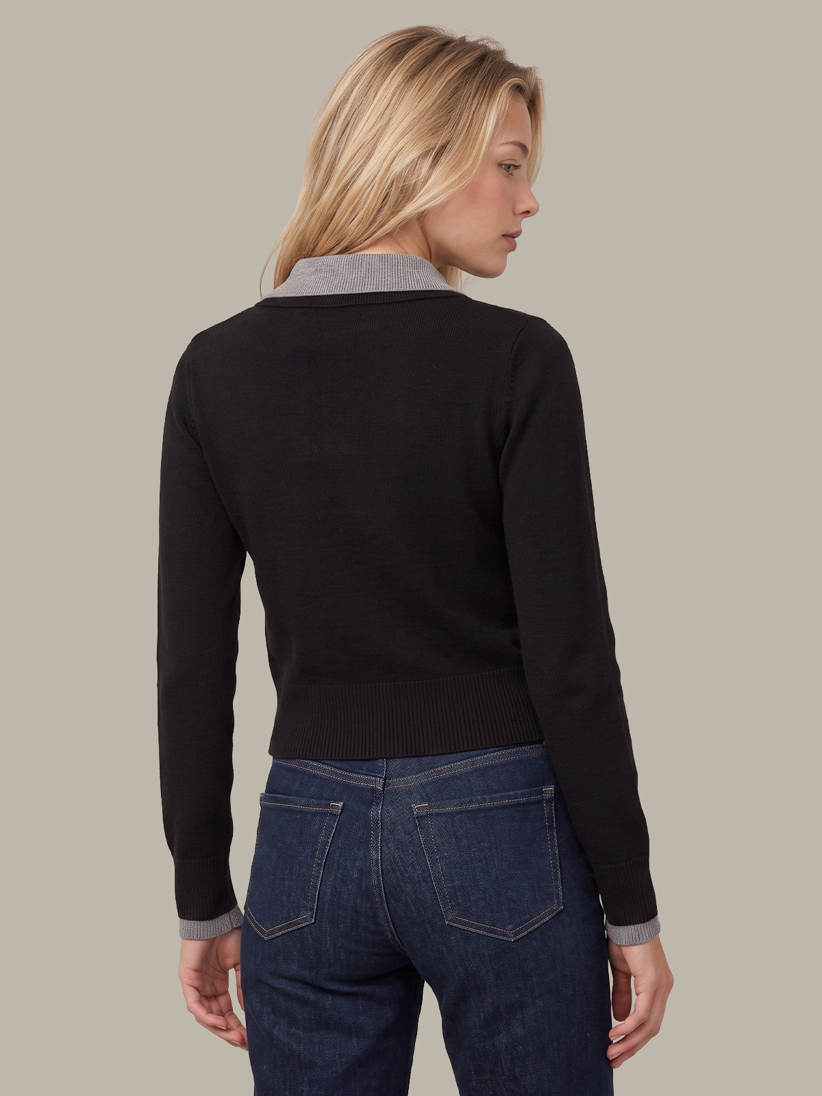 A woman shown from the back wearing a black long-sleeve knit sweater with a contrasting light gray collar and cuffs, paired with dark blue jeans, against a solid light gray background.