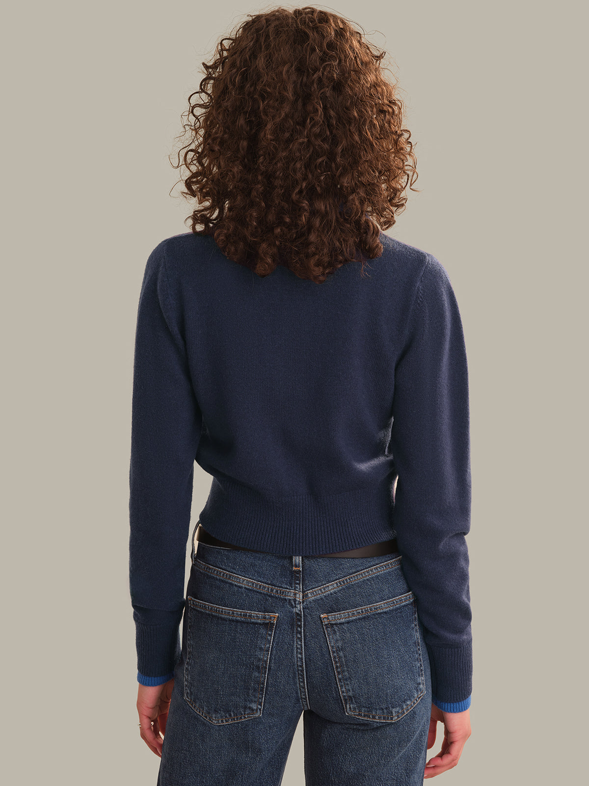 A person shown from the back wearing a navy blue long-sleeve knit cardigan and blue jeans against a plain warm gray background.