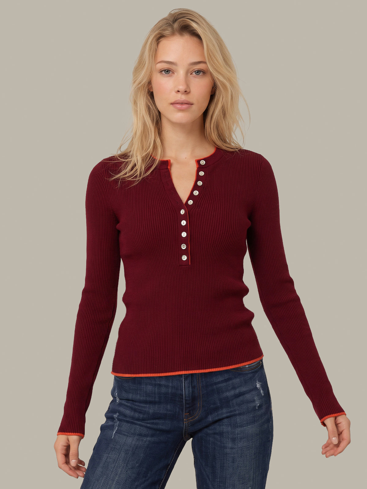 A woman modeling a ribbed long-sleeve henley sweater in deep burgundy with white buttons and orange trim, paired with blue jeans against a solid gray background.