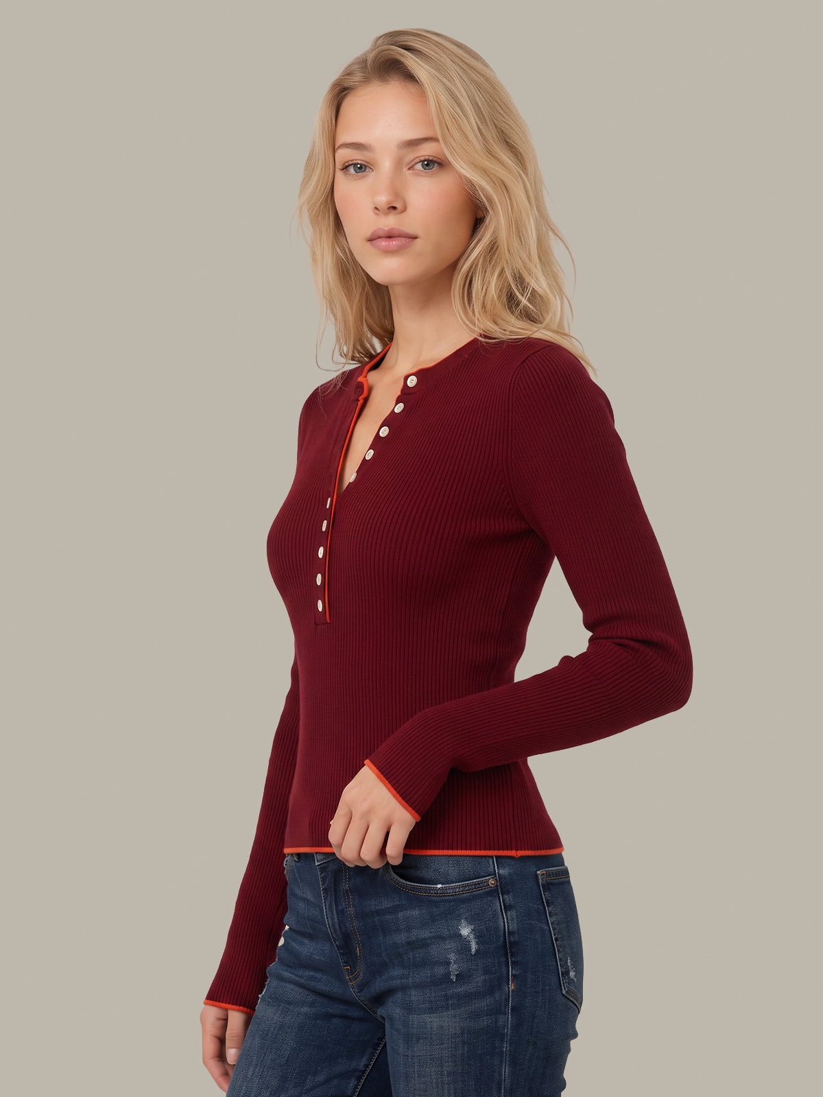 A woman wearing a fitted ribbed long-sleeve henley top in deep burgundy with decorative buttons down the front, paired with blue jeans, against a solid light gray studio background.