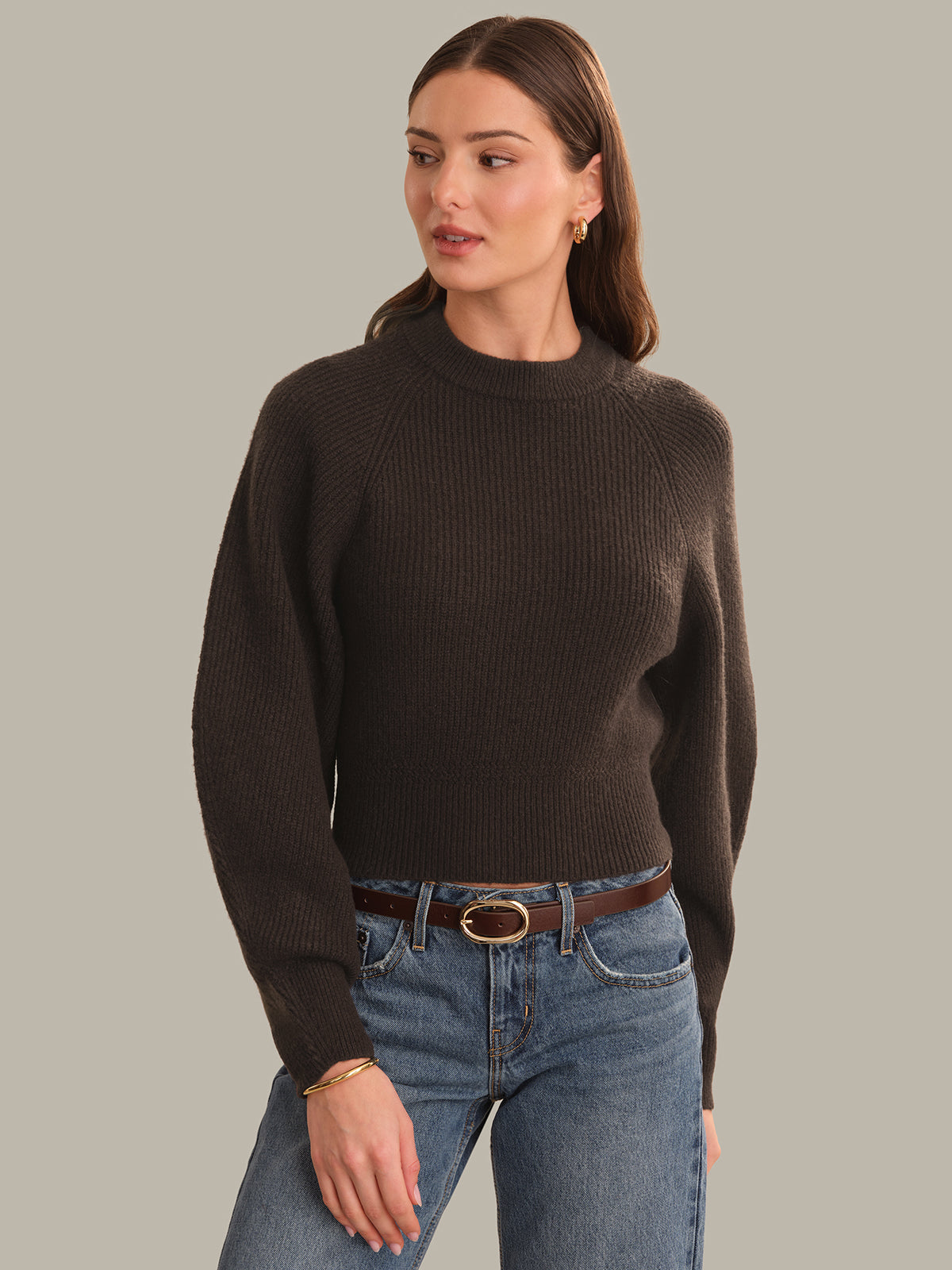 A woman modeling a dark brown ribbed knit mock neck sweater with raglan sleeves, paired with blue jeans and a brown belt, against a solid light gray background.