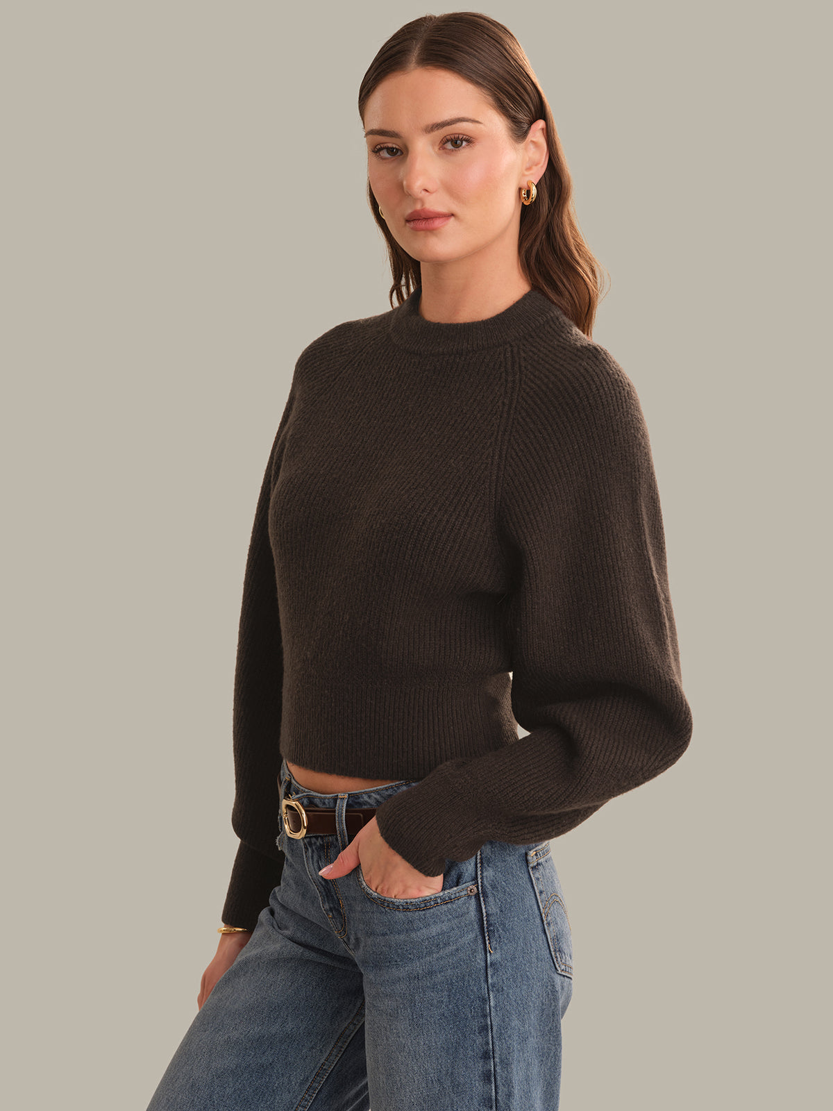 A woman modeling a dark brown ribbed knit mock neck sweater with long sleeves, paired with blue jeans, against a solid light gray background.