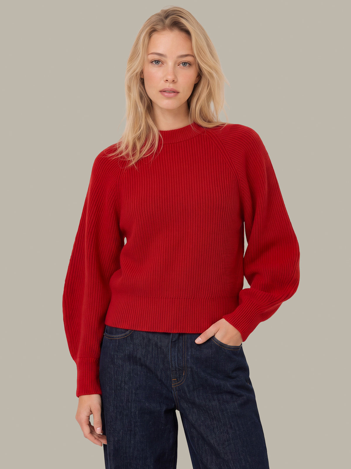 A woman wearing a vibrant red ribbed knit sweater with long raglan sleeves and dark blue jeans, standing against a solid neutral gray background.