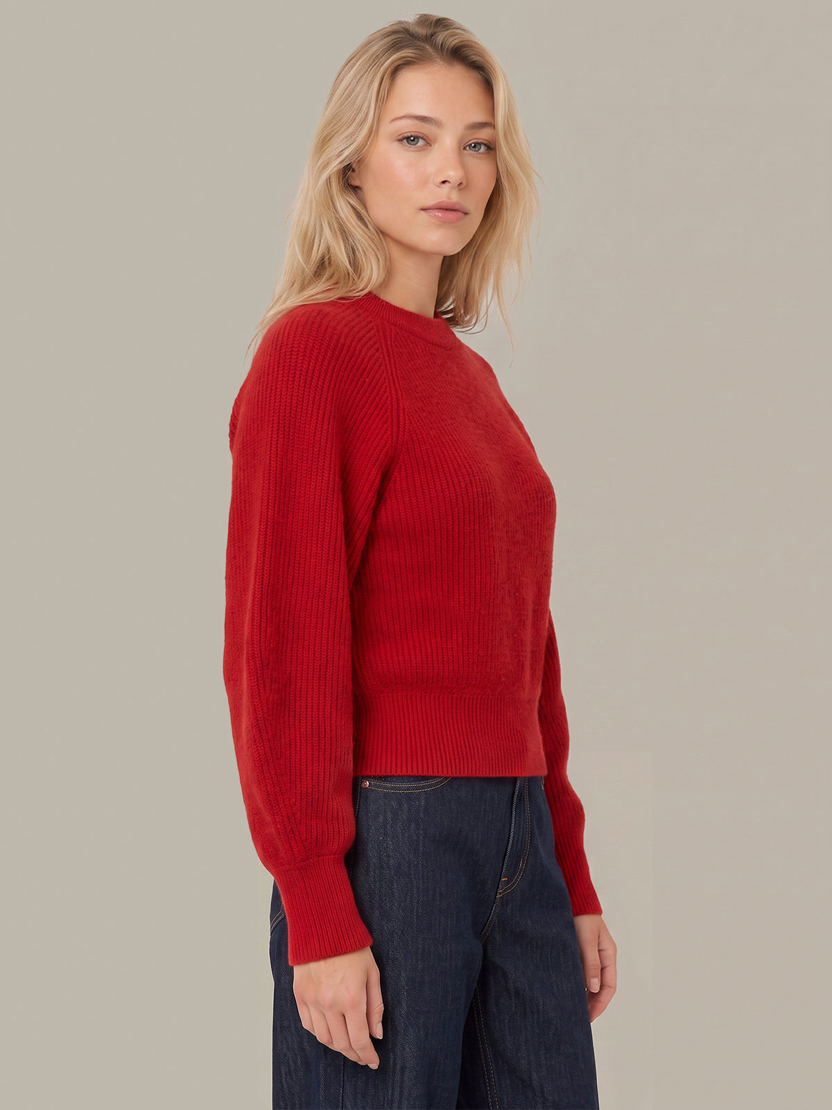 A woman wearing a red ribbed knit sweater and dark blue jeans, photographed against a solid light gray studio background.