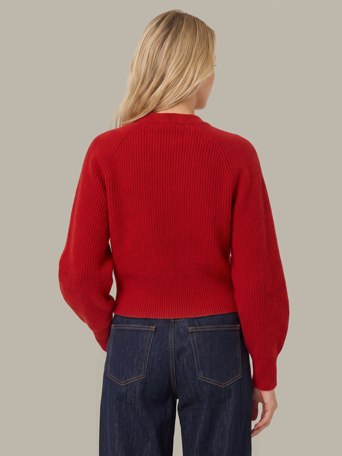 A woman viewed from the back wearing a bright red ribbed knit sweater and dark blue jeans, set against a solid light gray background.