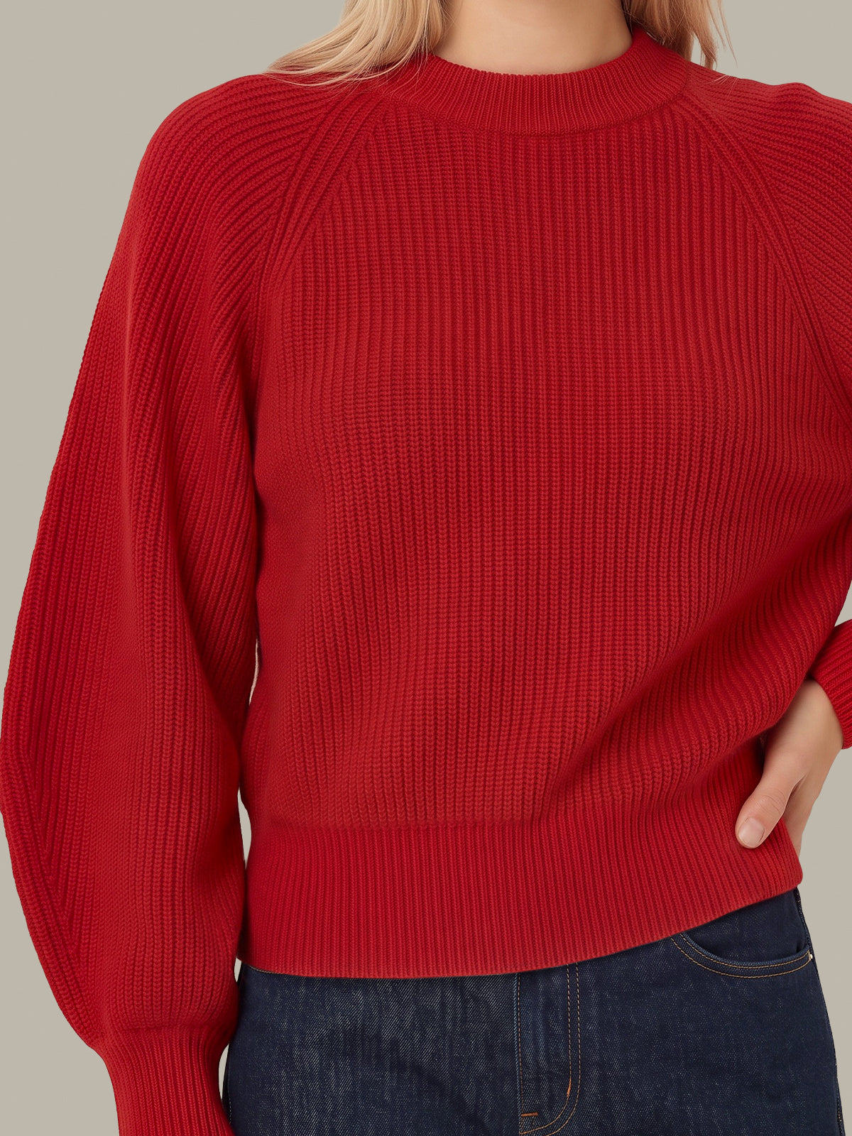 A close-up of a person wearing a red ribbed knit sweater with balloon sleeves and a crew neck, paired with dark blue jeans against a plain beige background.