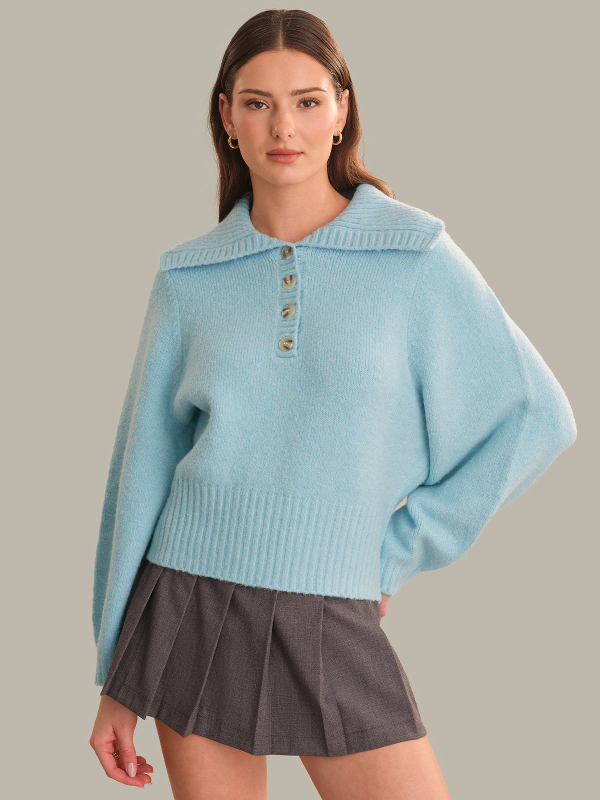 A woman modeling a light blue knit sweater with a wide collar and three front buttons, paired with a dark gray pleated mini skirt, against a solid beige background.