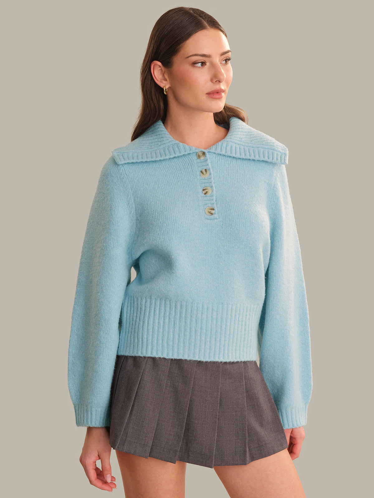 A woman modeling a light blue knit sweater with a wide collar and three decorative buttons at the neckline, paired with a dark gray pleated mini skirt, against a solid taupe background.