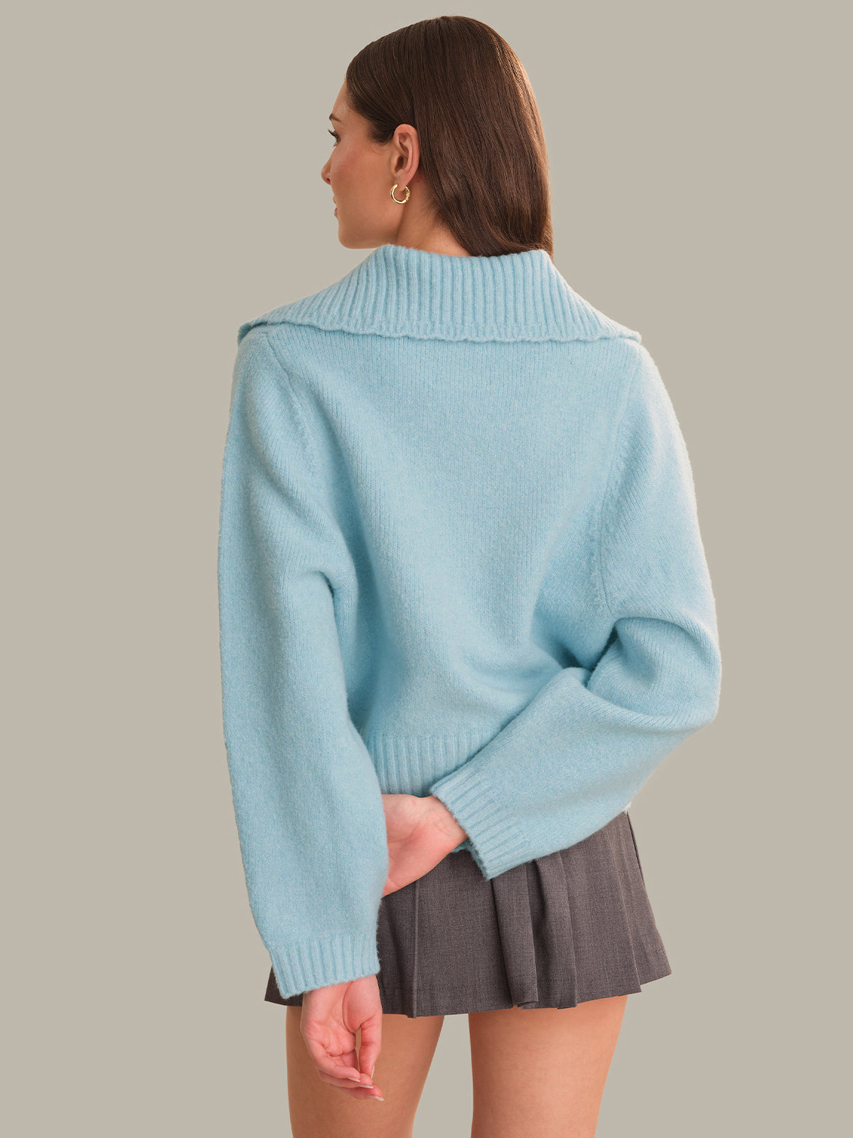 A woman shown from the back wearing a light blue knit sweater with a wide ribbed collar and a dark gray pleated mini skirt, against a solid beige background.