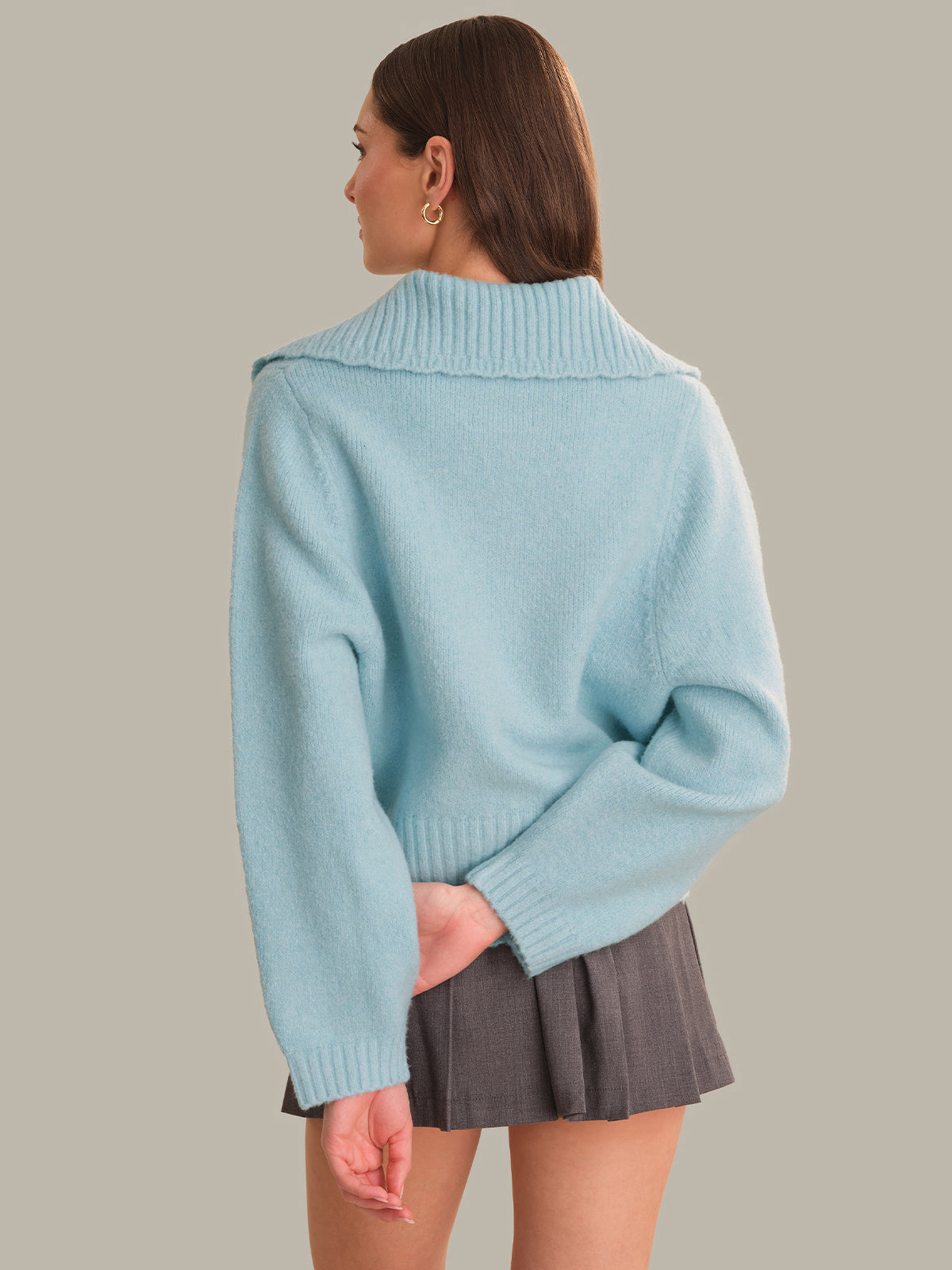 A woman shown from the back wearing a light blue knit sweater with a wide ribbed collar and a gray pleated mini skirt, against a solid warm gray background.