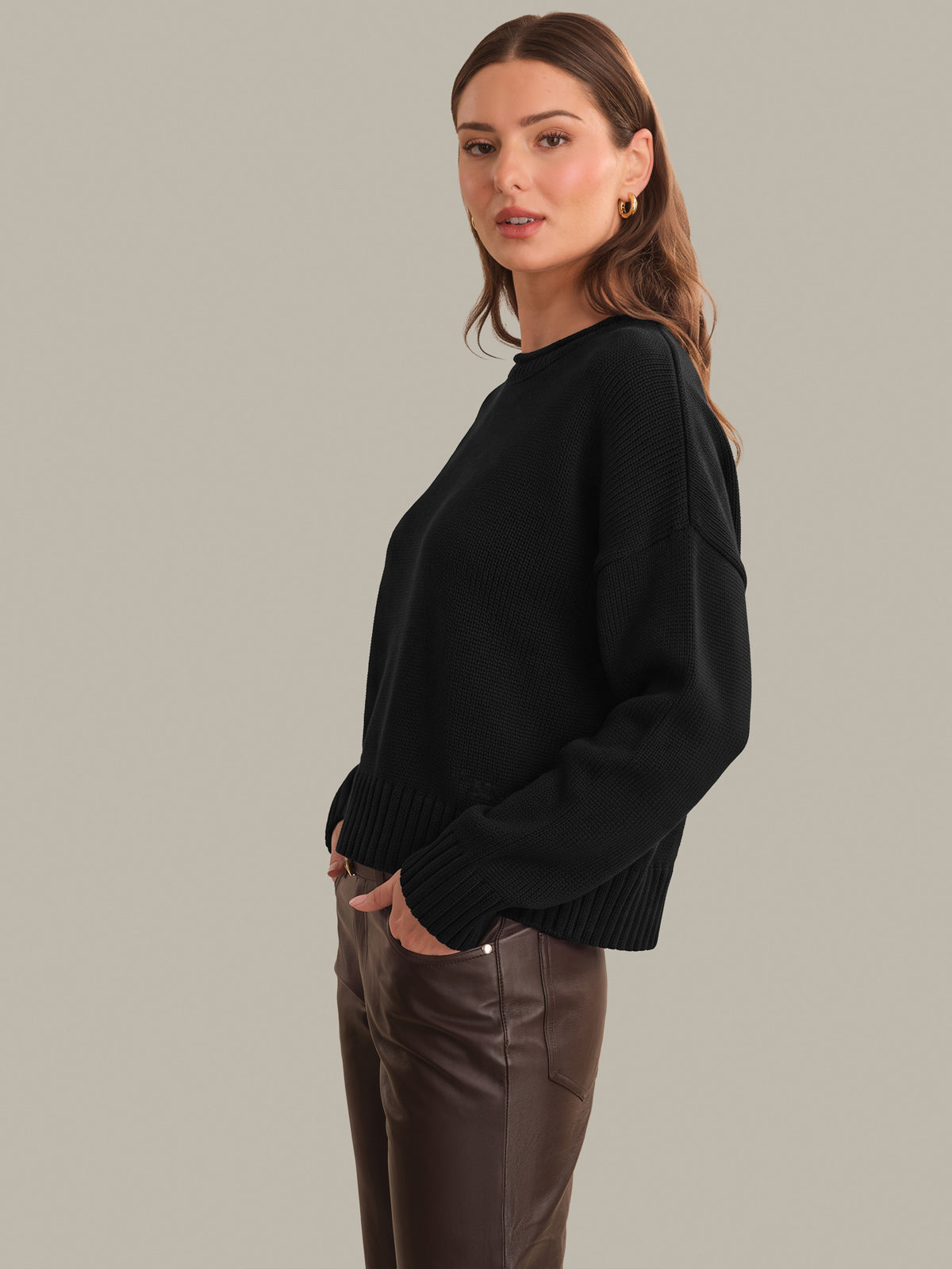 A woman modeling a black knit crewneck sweater with ribbed cuffs and hem, paired with brown pants, against a solid light gray background.