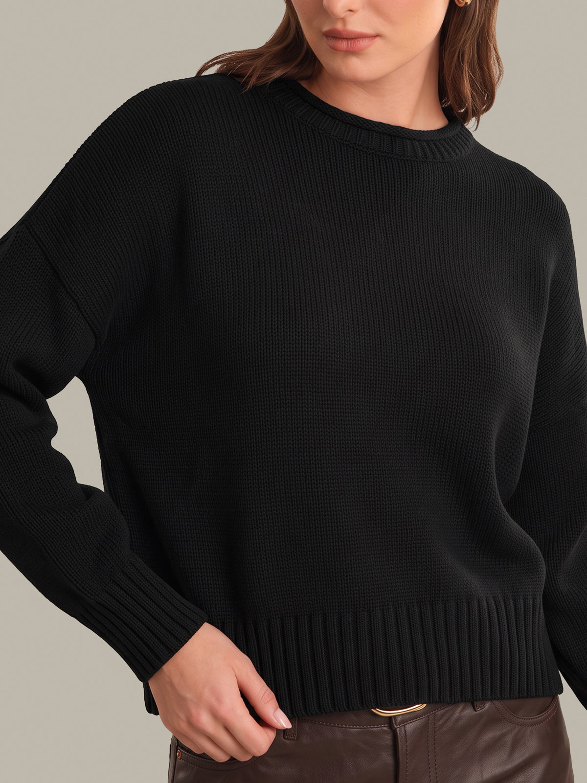 A close-up of a woman wearing a black ribbed knit sweater with a crew neck and dropped shoulders, paired with brown leather pants, against a solid light gray background.