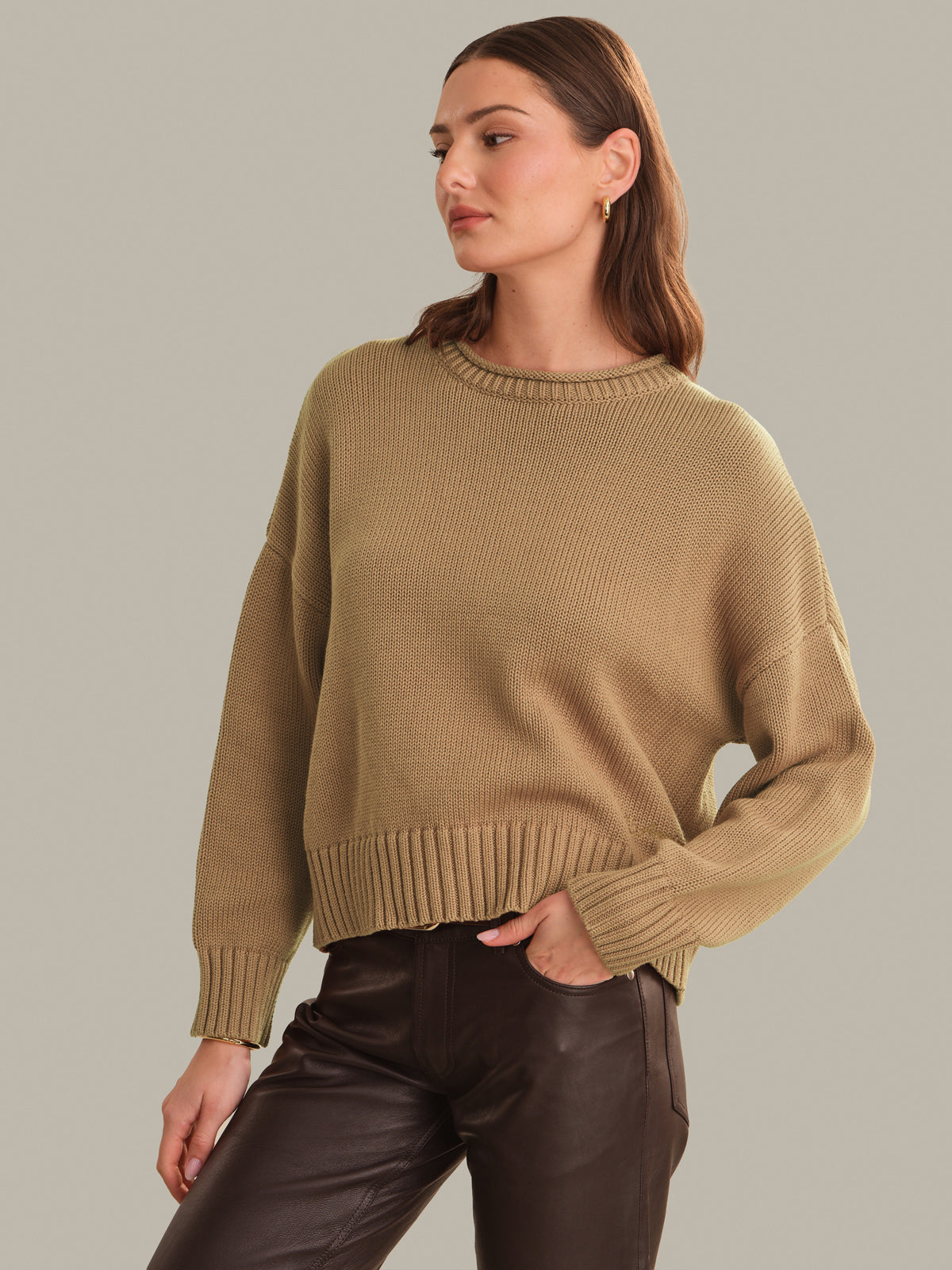 A woman wearing a cropped, ribbed knit sweater in a warm camel tan color, paired with dark brown faux-leather pants, against a solid light gray studio background.