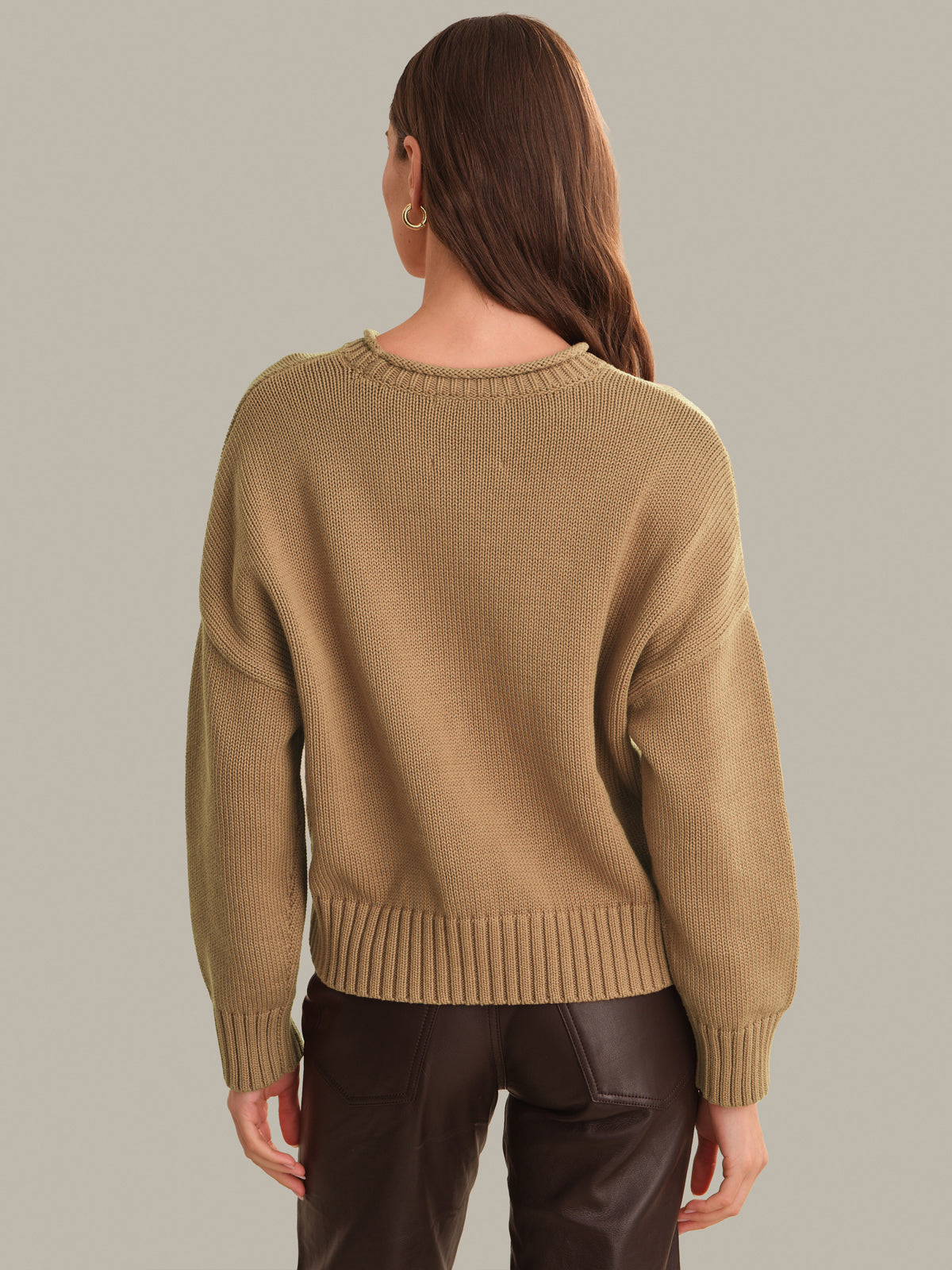 A woman viewed from the back wearing a camel-colored ribbed knit sweater and dark brown pants, set against a solid light gray studio background.