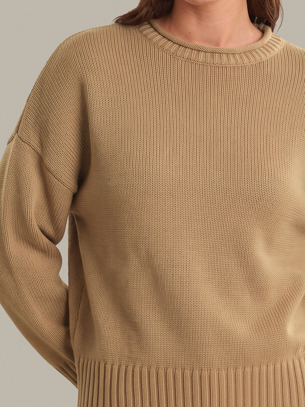 A close-up of a person wearing a textured knit crewneck sweater in a warm camel tan color, showing the ribbed collar, sleeve seams, and ribbed hem against a neutral light gray background.