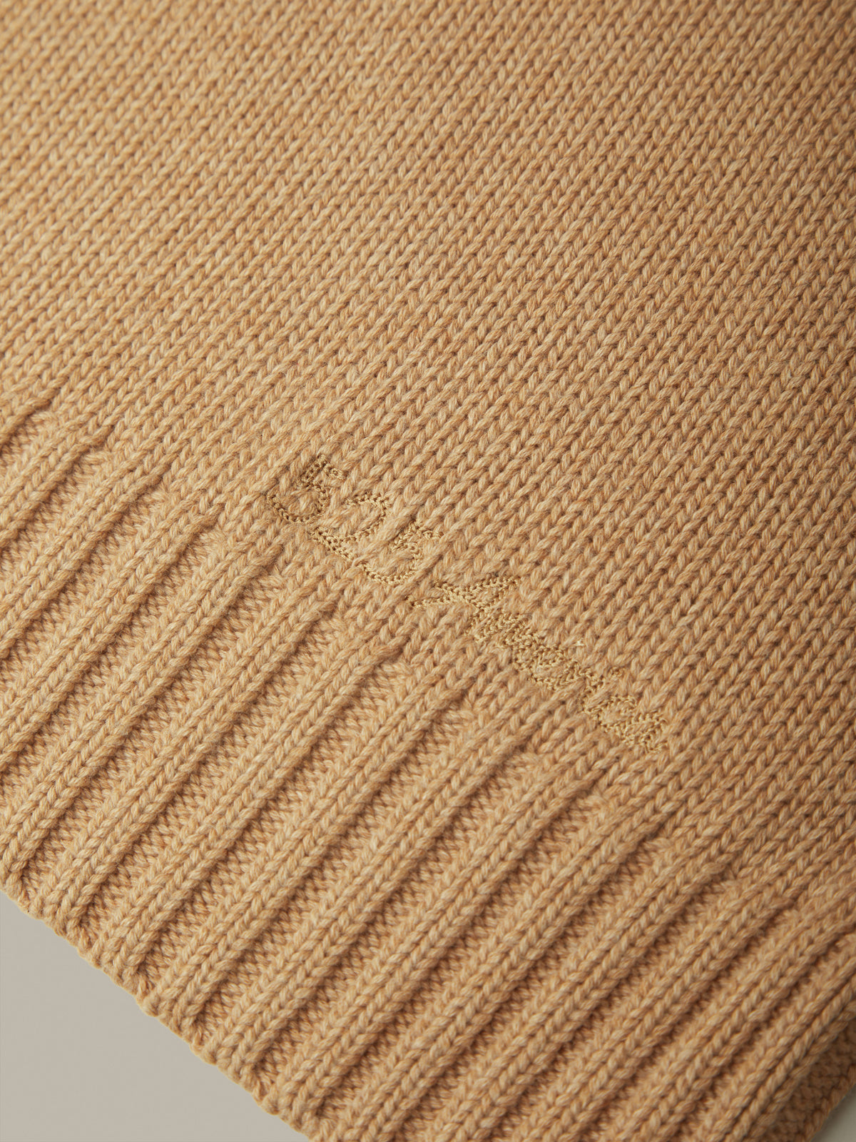 Close-up of a ribbed knit sweater in a warm camel beige color, showing the fine knit texture and a subtle tonal embroidered logo.