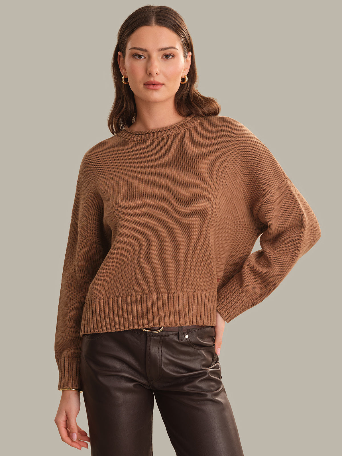 A woman modeling a cropped, long-sleeve ribbed knit sweater in a warm camel brown color, paired with black faux-leather pants, against a solid light gray background.